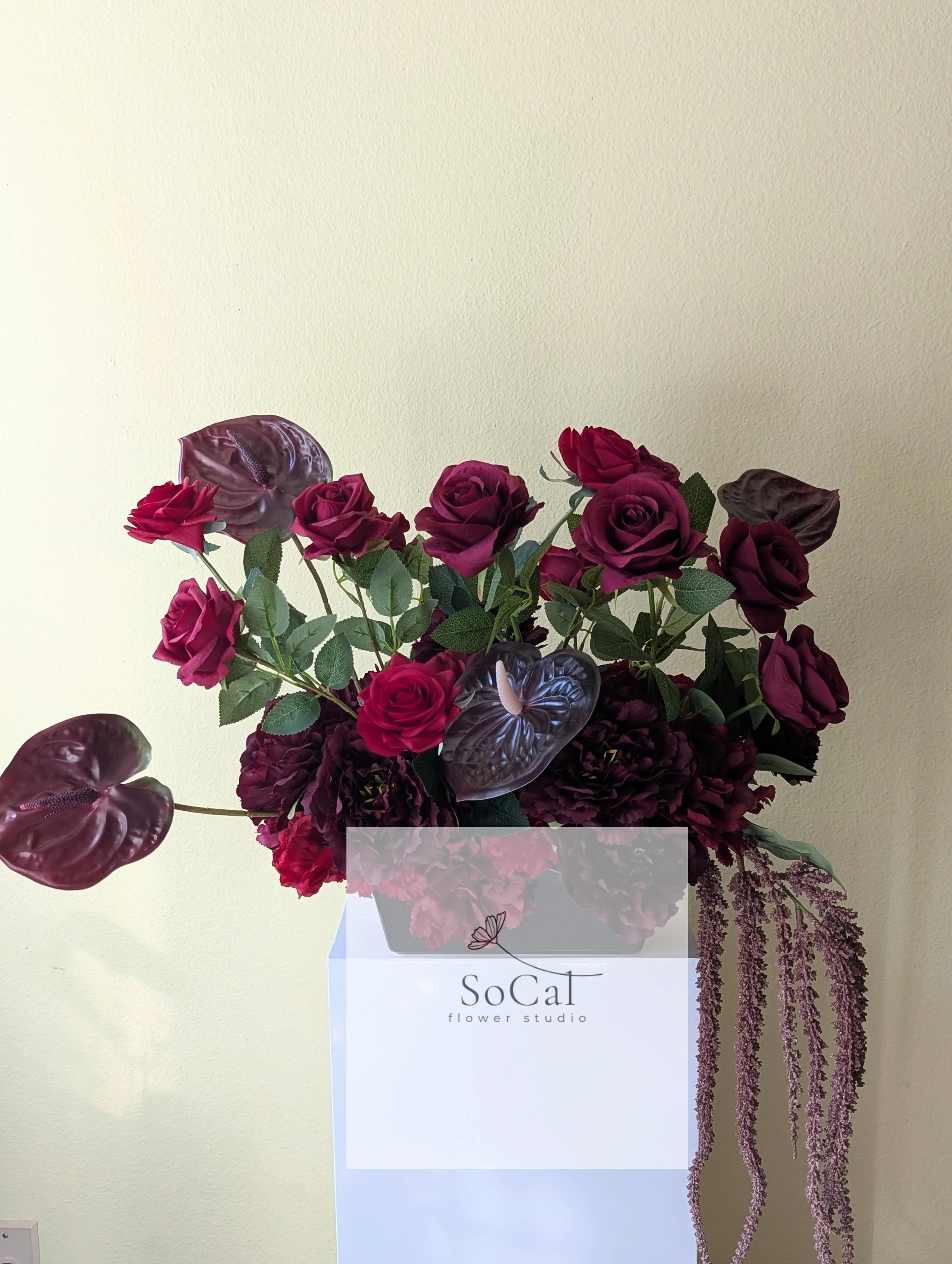 Bouquet of burgundy roses, anthuriums, and pinkish hanging amaranthus in a white stand with 'SoCal flower studio' logo.