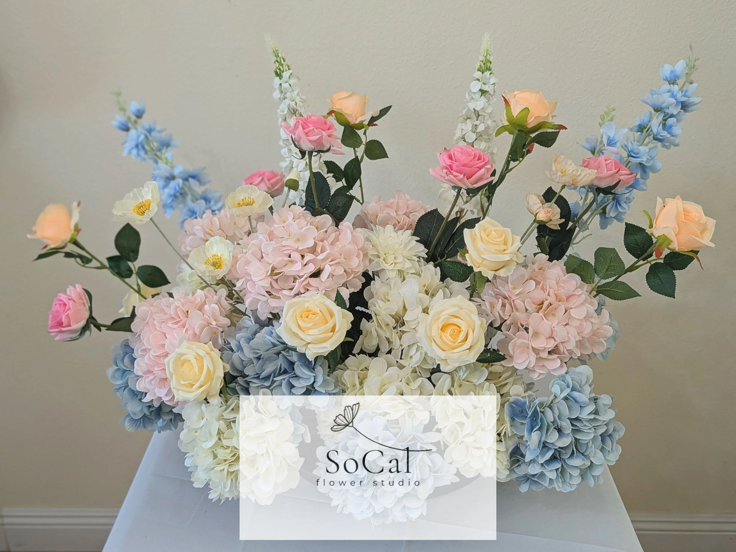A floral arrangement with pink, cream, blue, and white roses, hydrangeas, and other flowers in a black vase on a white table against a beige wall.