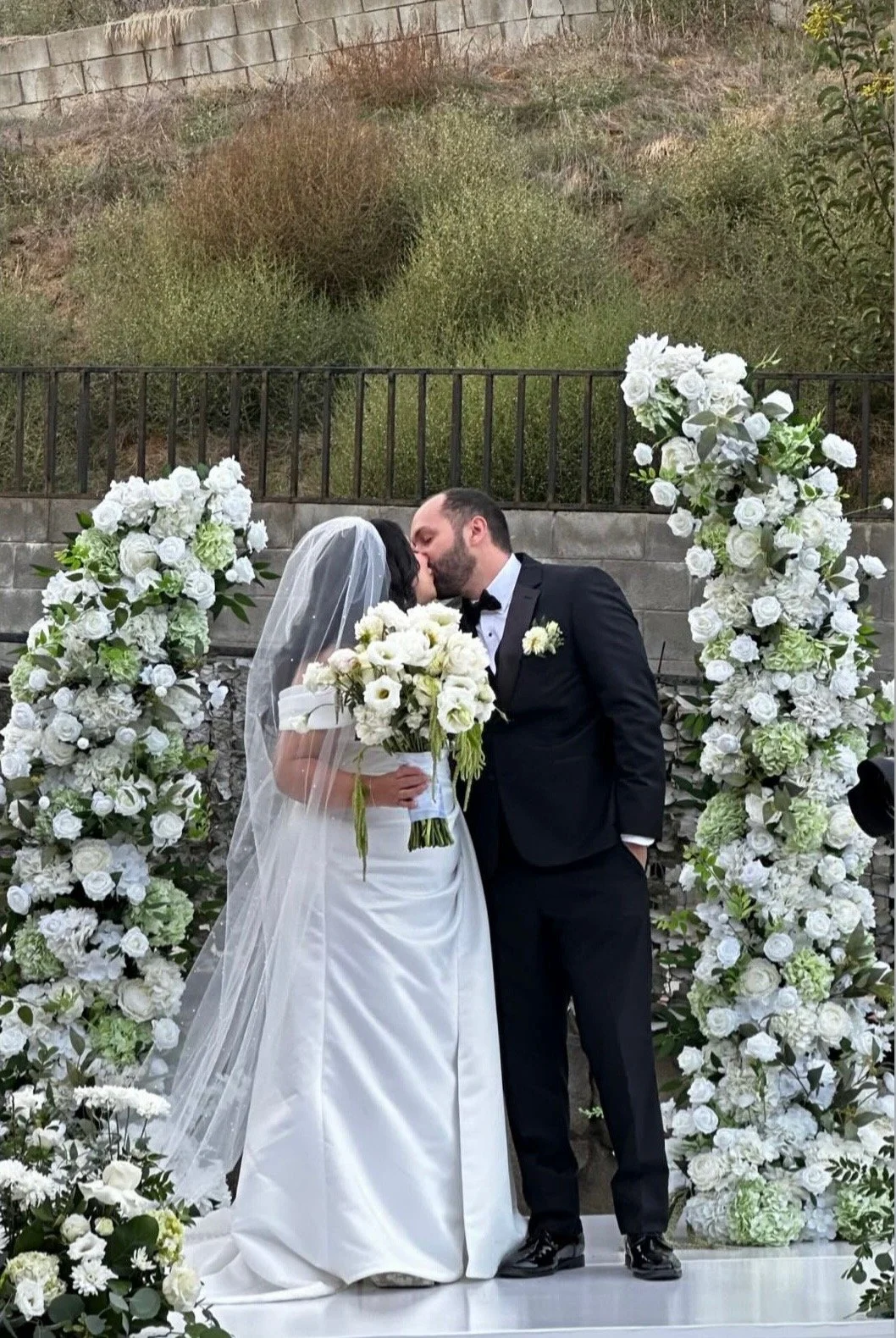 A bride and groom share a kiss at their wedding, standing between two tall arrangements of white roses and greenery. The bride wears a white gown and veil, holding a bouquet of white flowers, while the groom is dressed in a black tuxedo with a bow ti