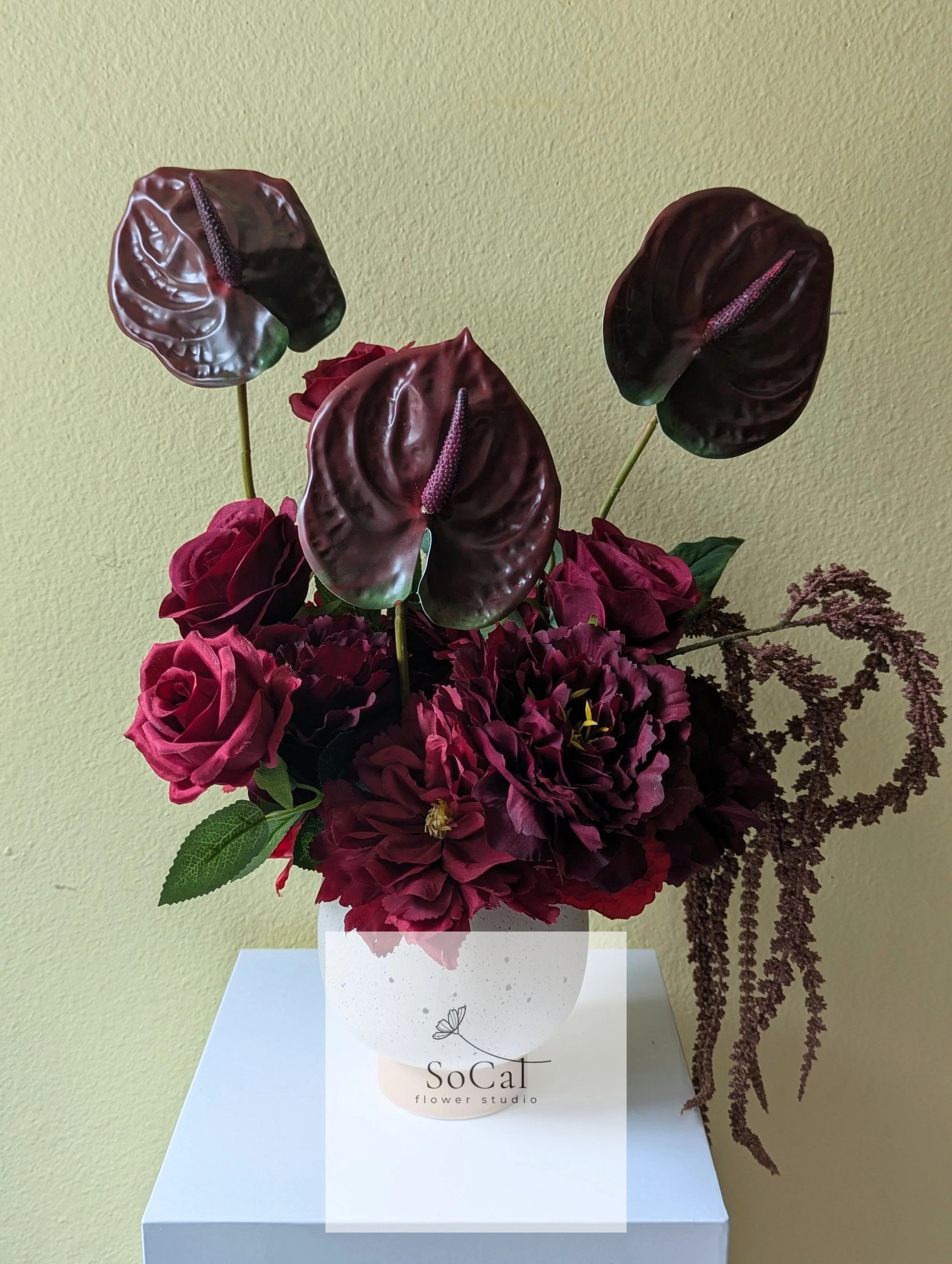 Dark red anthurium flowers and burgundy roses in a white flower vase on a white surface against a light green wall.