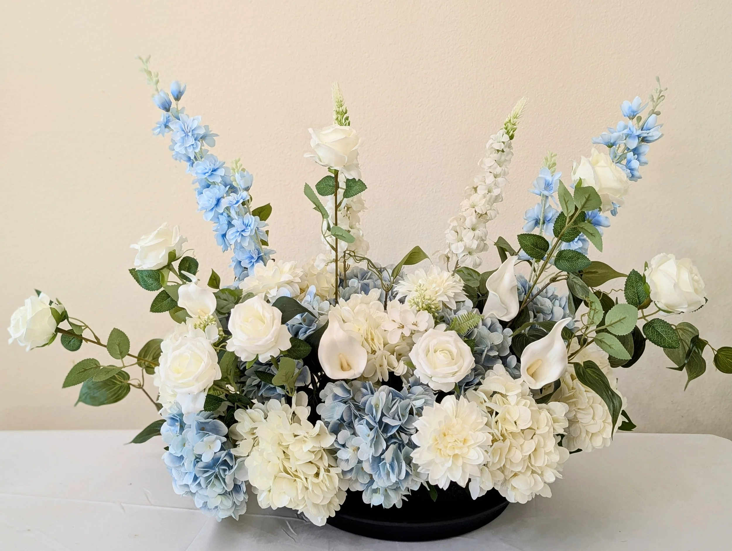 Arrangement of white roses, calla lilies, and blue hydrangeas with green leaves in a black vase.