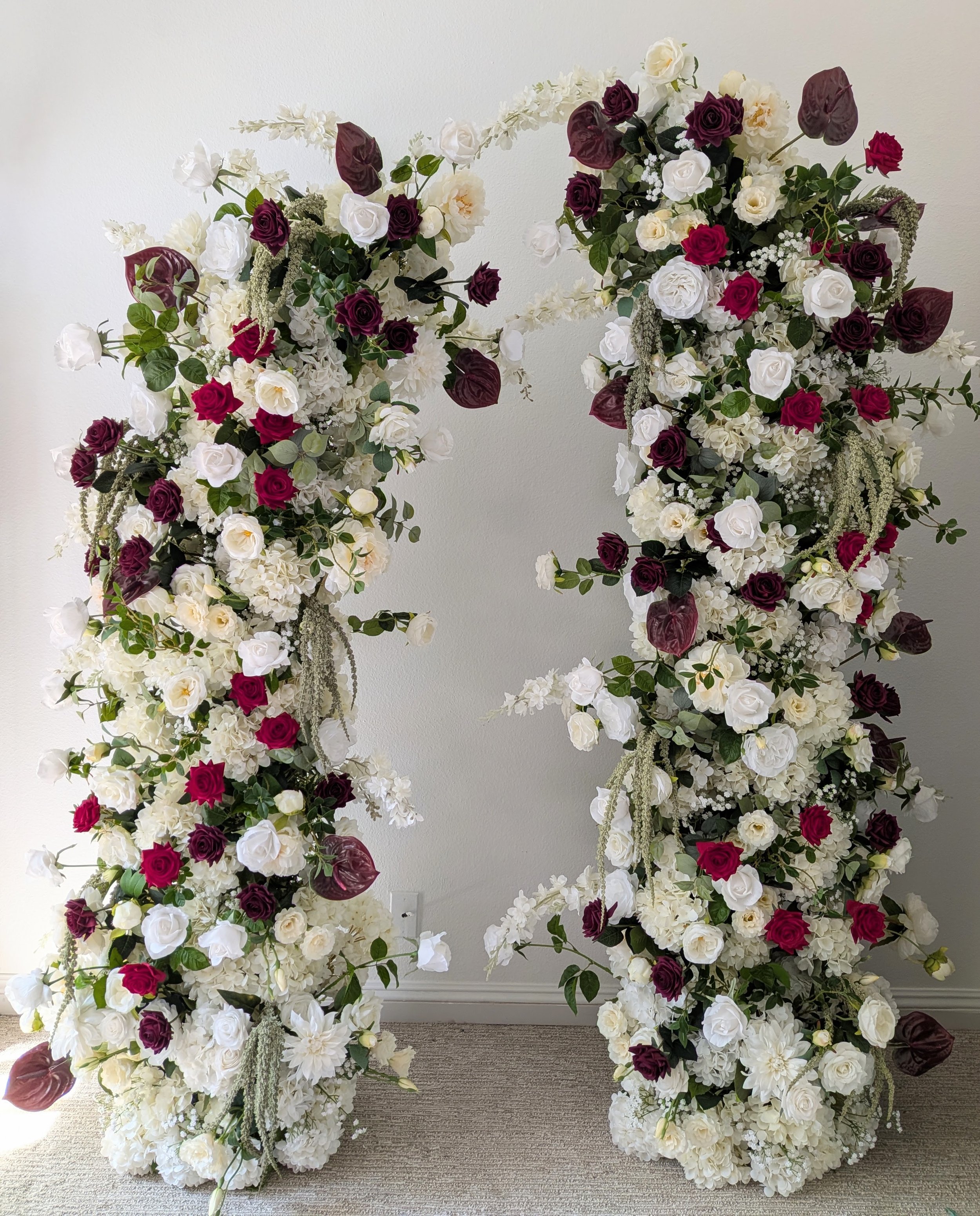 A floral arrangement with white, dark purple, and red roses, along with white hydrangeas and green foliage, standing against a plain off-white wall.