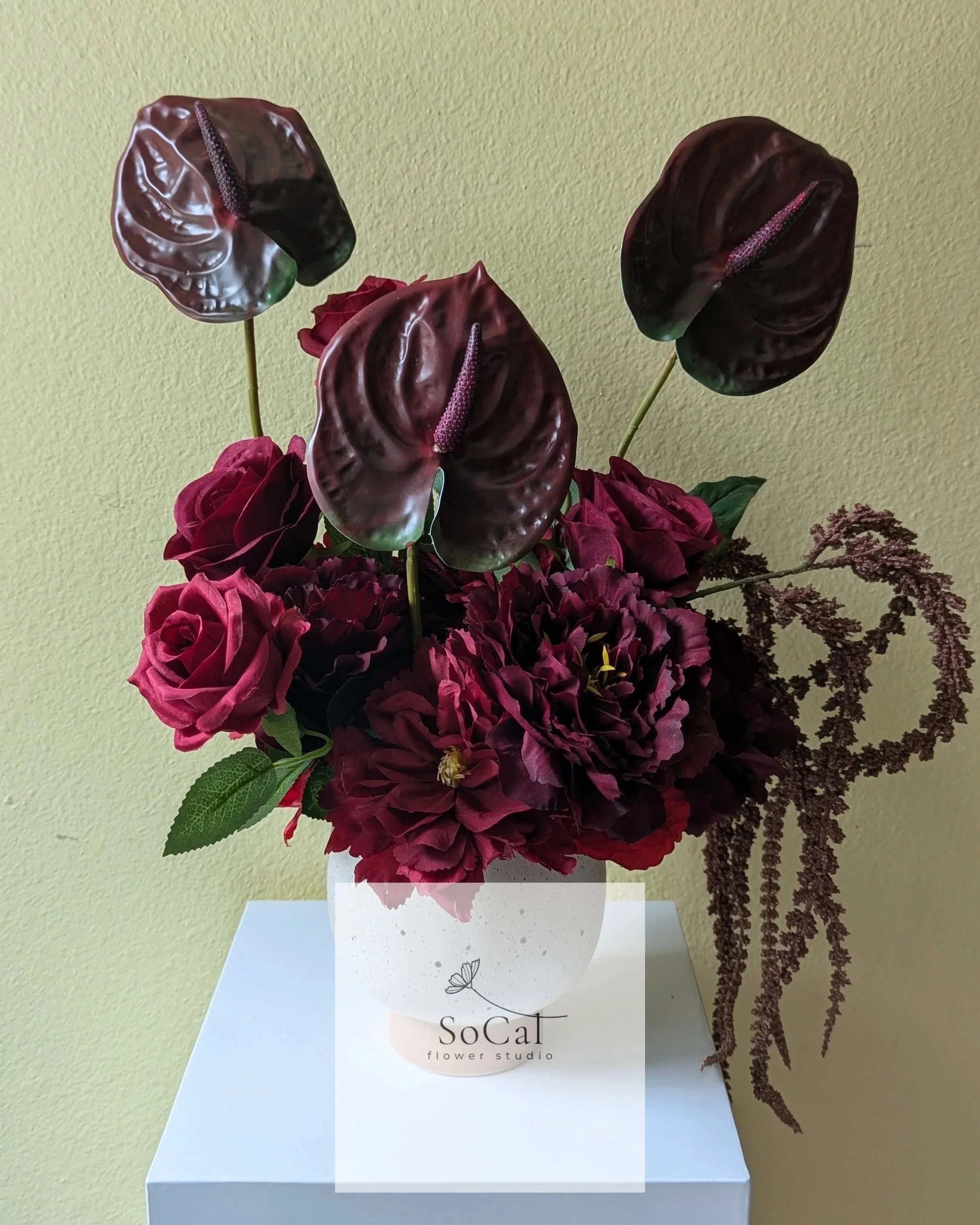 Arrangement of dark red hydrangeas, black calla lilies, and purple-leaved plants in a white square vase on a plain wall background.