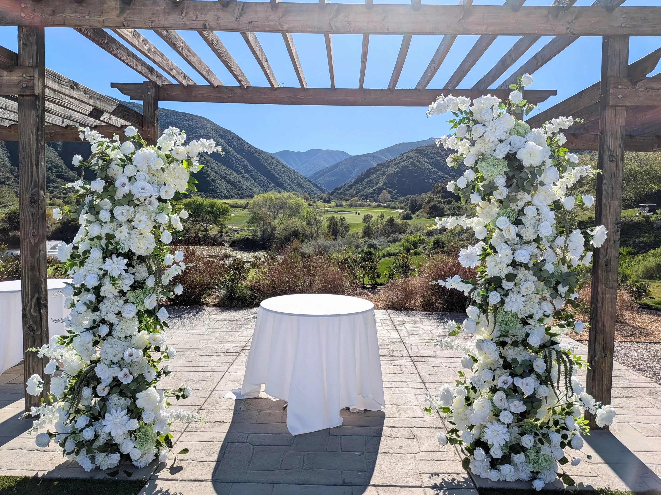 A wedding arch decorated with white flowers on a patio overlooking a lush green valley and mountains under a clear blue sky.