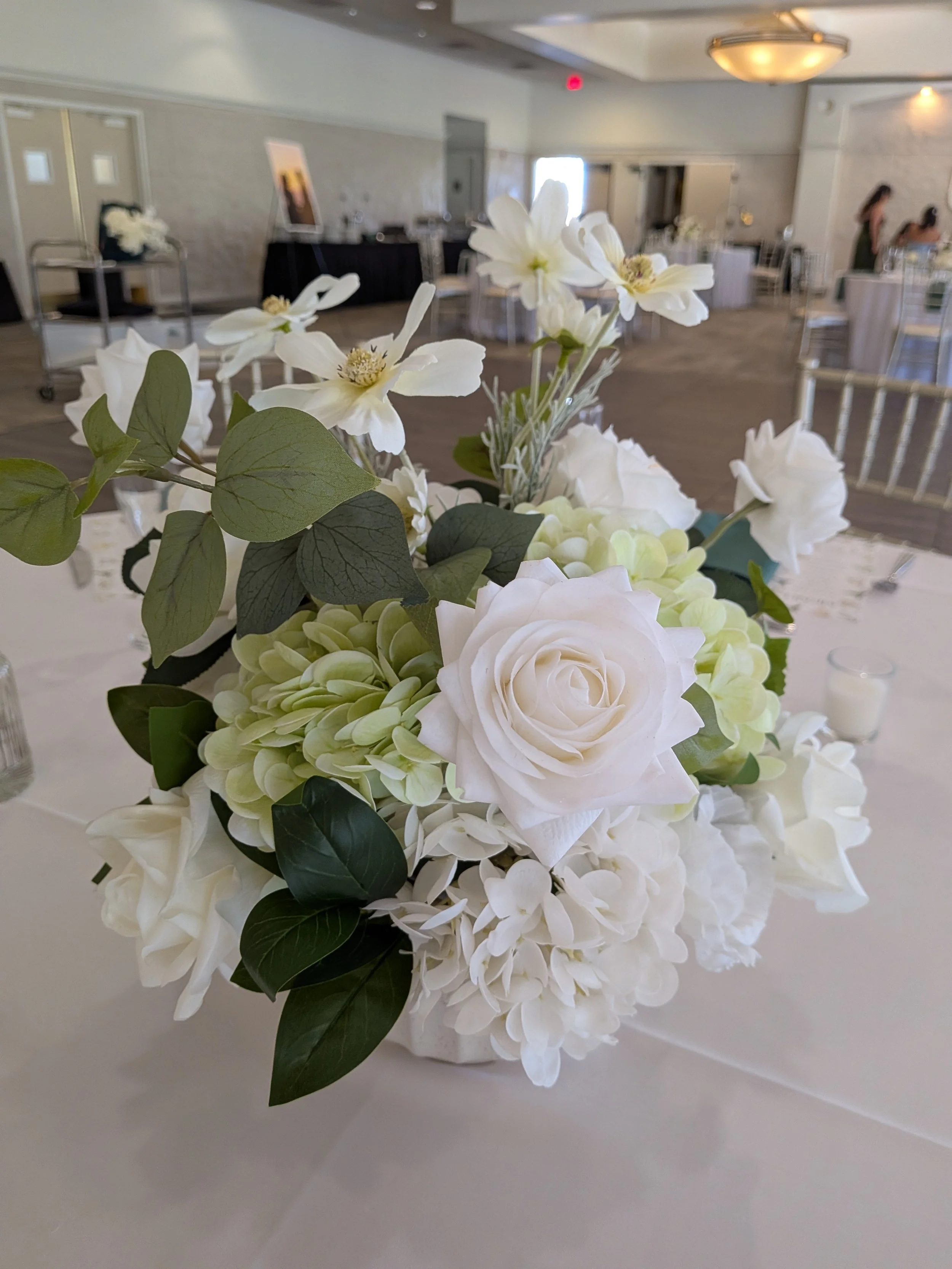 A white floral centerpiece featuring roses, hydrangeas, and greenery on a table in an event space.
