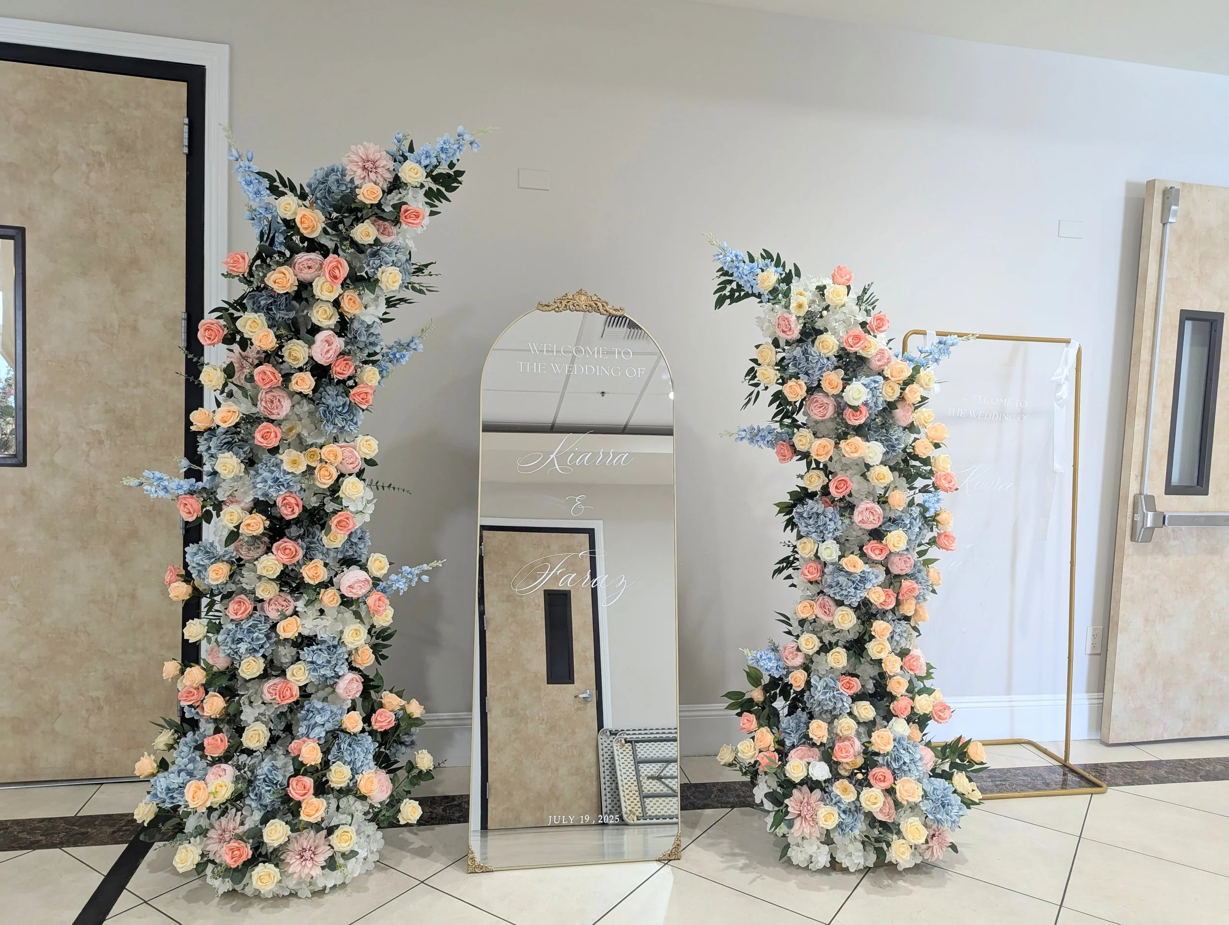 Two large floral archways with pink, peach, white, and blue flowers, flanking a tall mirror with gold trim and Eiffel Tower decoration, at a wedding venue.