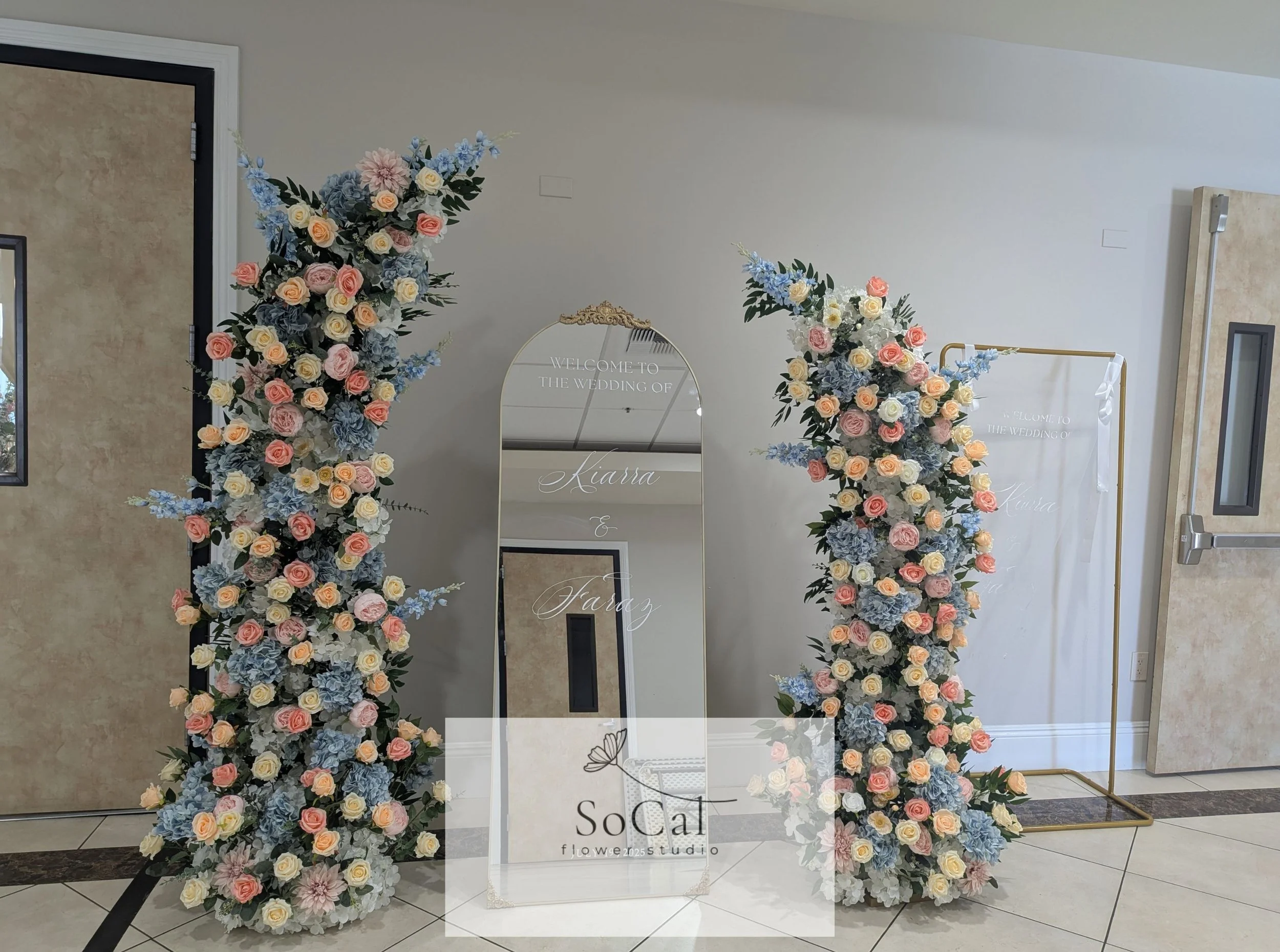 Floral wedding arch with pink, cream, and blue flowers, a mirror with wedding text, and a stand with wedding signs.