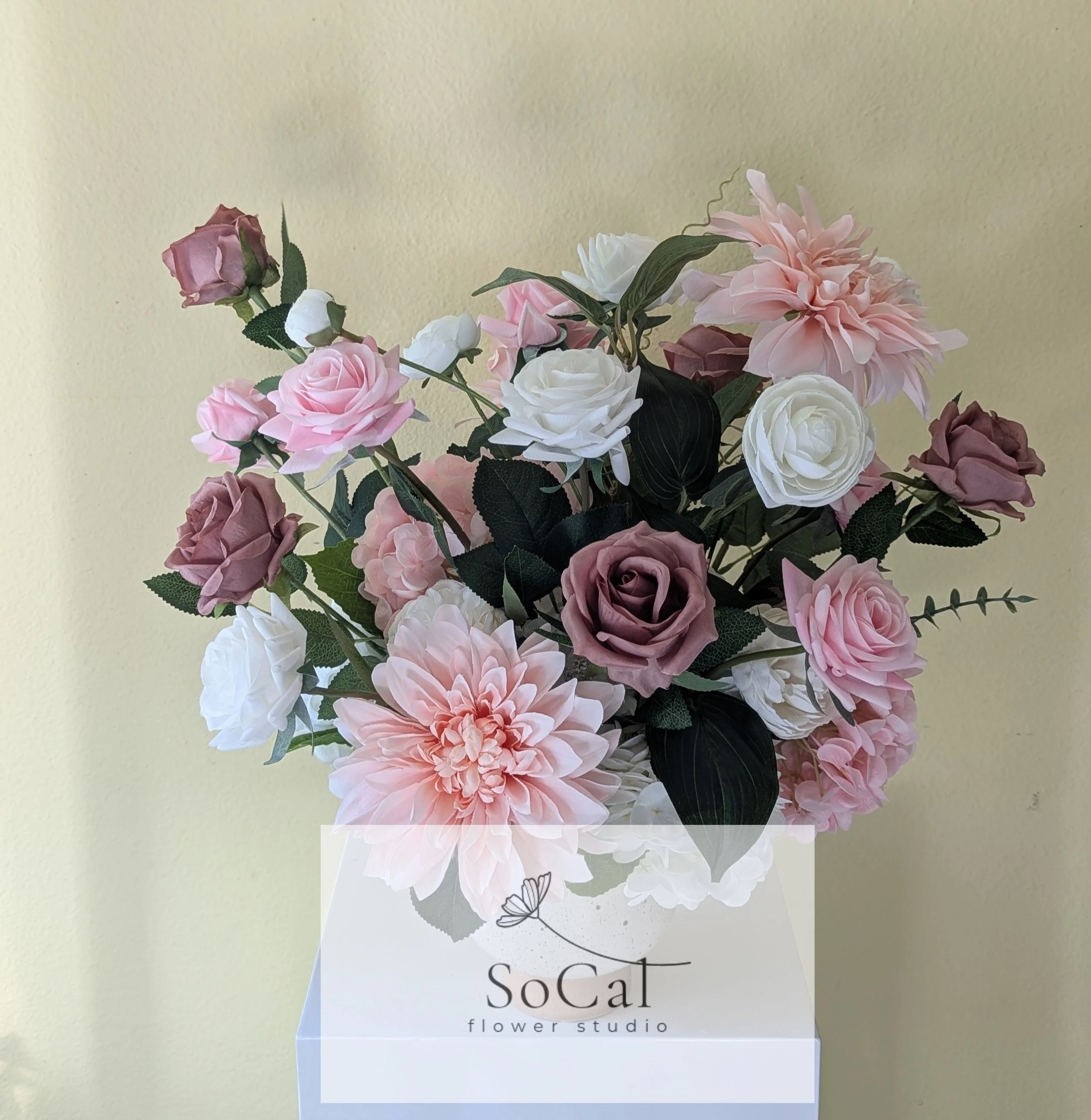 A bouquet of pink, white, and purple artificial flowers with green leaves in a white vase labeled 'SoCat flower studio' against a light yellow wall.