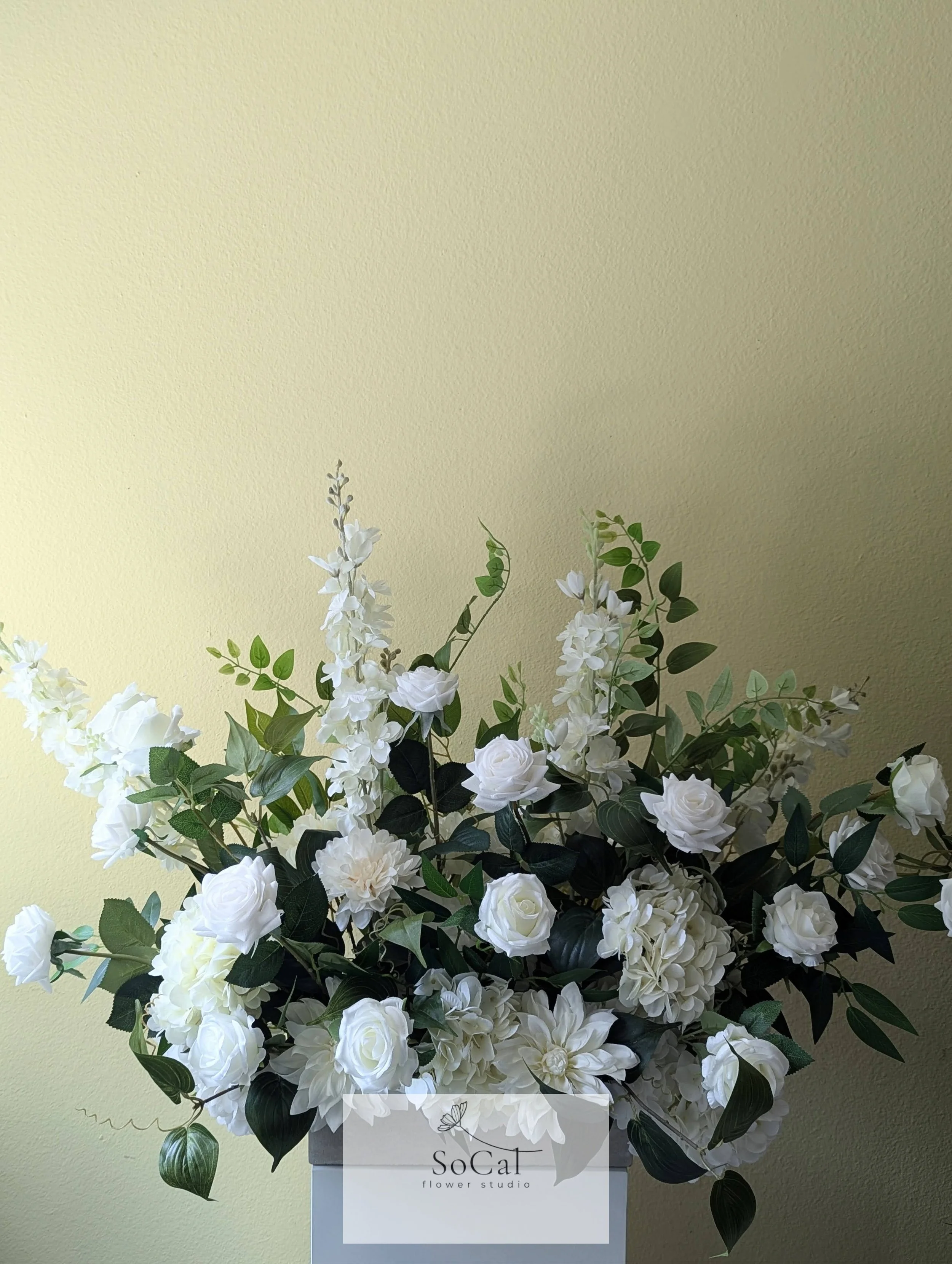 A floral arrangement with white roses, dahlias, and other white flowers, accented with green leaves, in a white box labeled 'SoCal flower studio' against a light yellow wall.