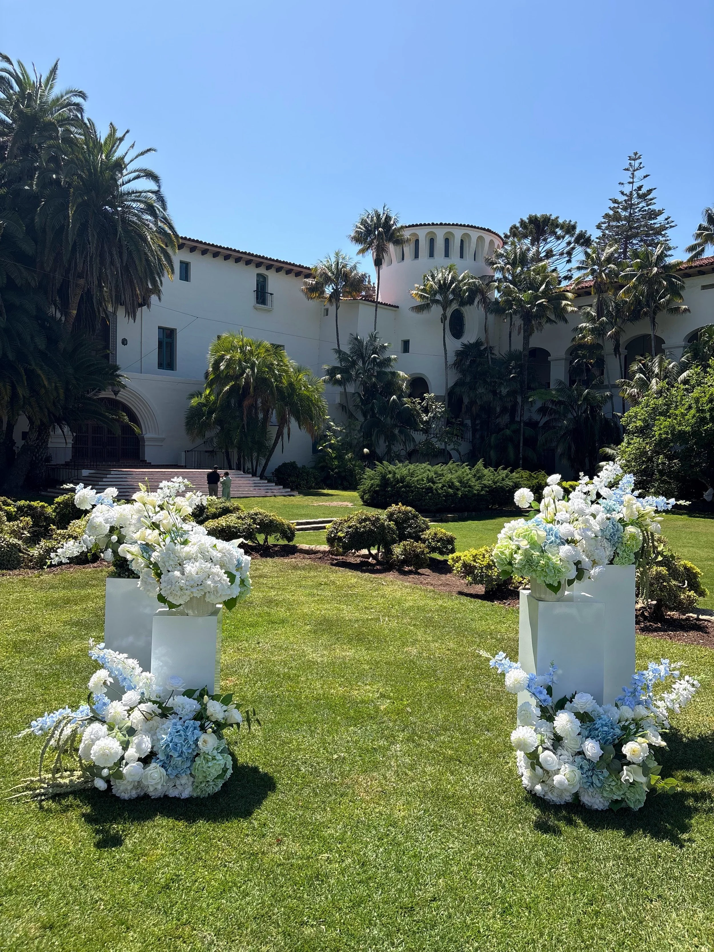 White flowers arranged on white stands are in the foreground on a lush green lawn, with a large white building surrounded by tall palm trees and other greenery in the background under a clear blue sky.