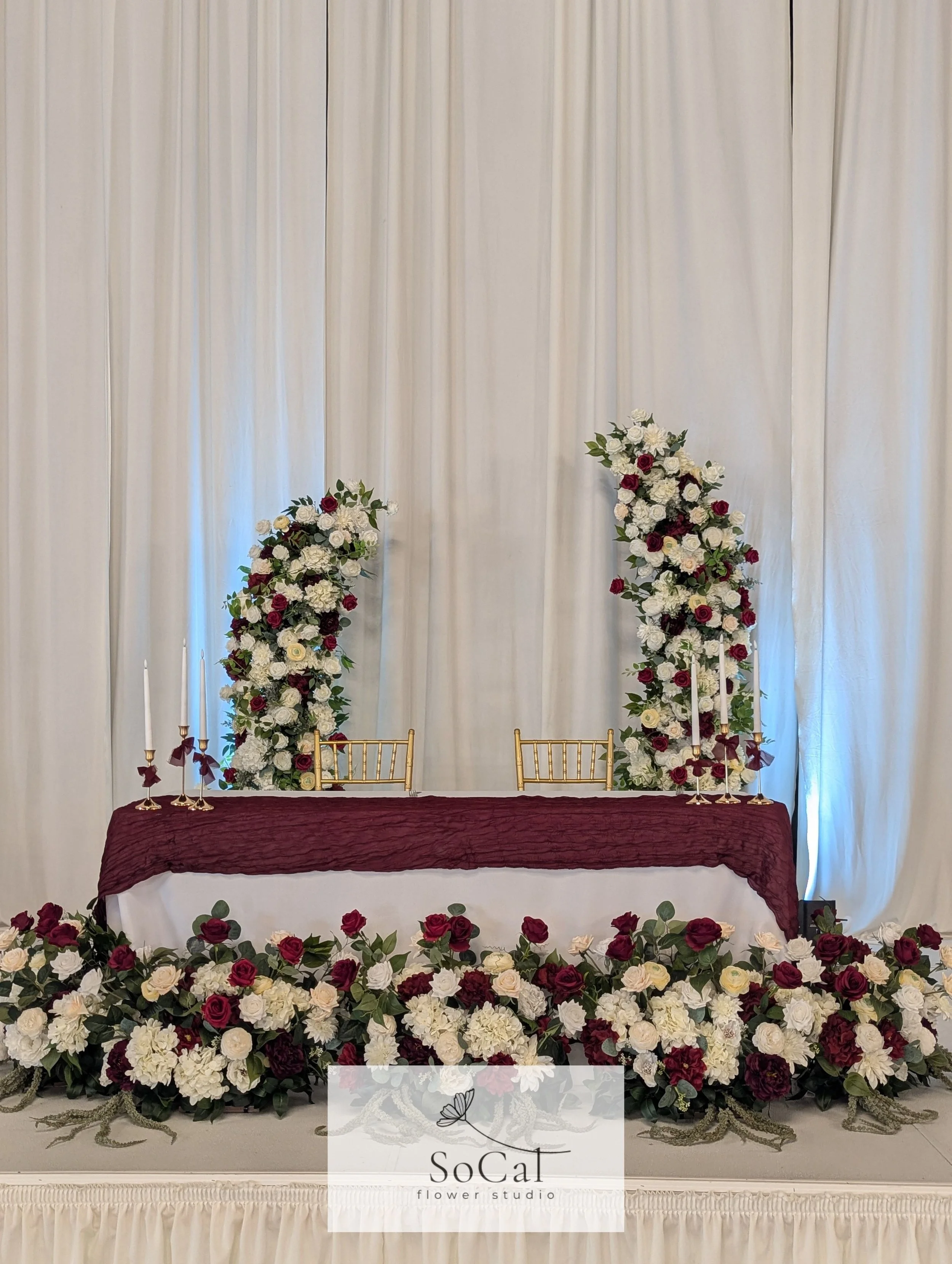 Wedding ceremony backdrop with white curtains, floral arrangements of white and dark red roses, and gold chairs behind a table with a burgundy tablecloth and floral decorations at the front, featuring the SoCal Flower Studio logo.
