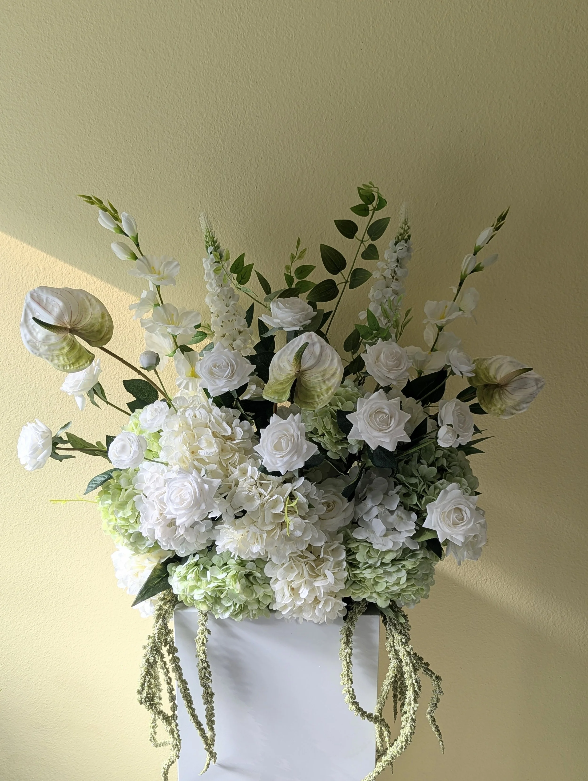 Arrangement of white flowers including roses, hydrangeas, and other blooms in a black vase on a white pedestal