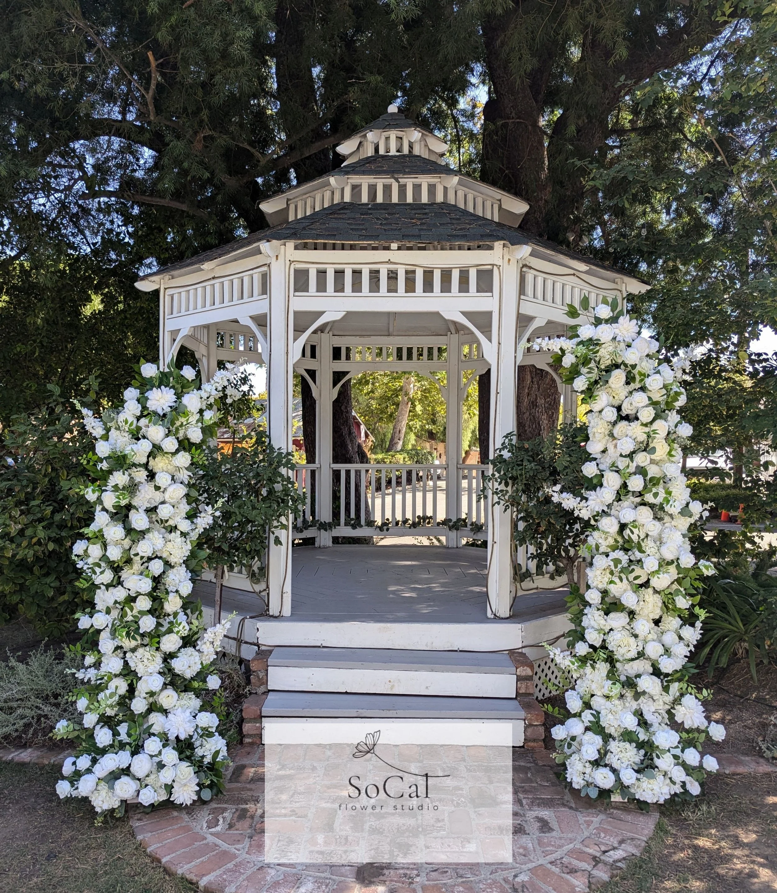 A white wooden gazebo decorated with large floral arrangements of white roses and other white flowers, situated outdoors with trees in the background.
