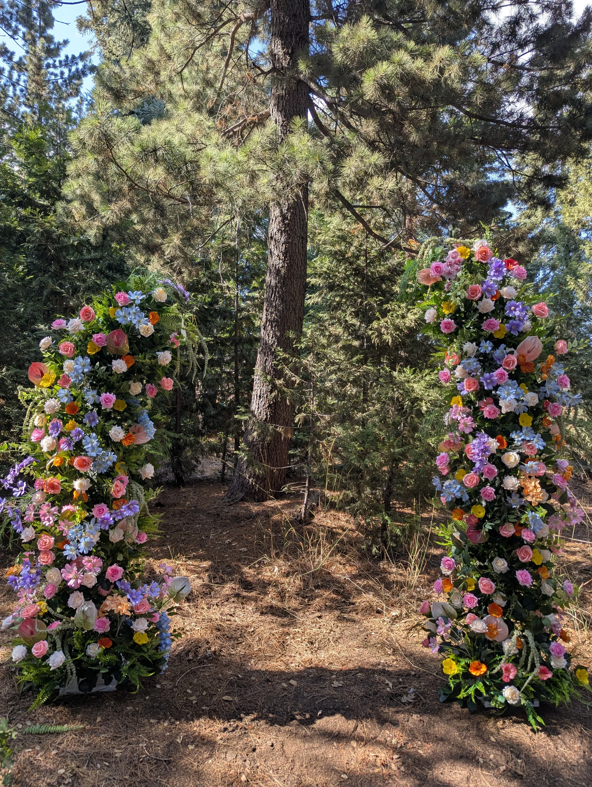 A floral archway made of colorful flowers such as roses, daisies, and other blossoms, set in a wooded outdoor area with trees and sunlight filtering through the branches.