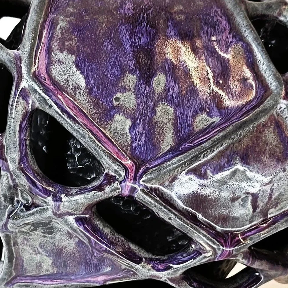 Mirsini Roumelioti. Mirsini Ceramics. Armor. Hugs. A metallic sculpture resembling a geometric face with purple and silver hues, featuring open spaces and intricate details.