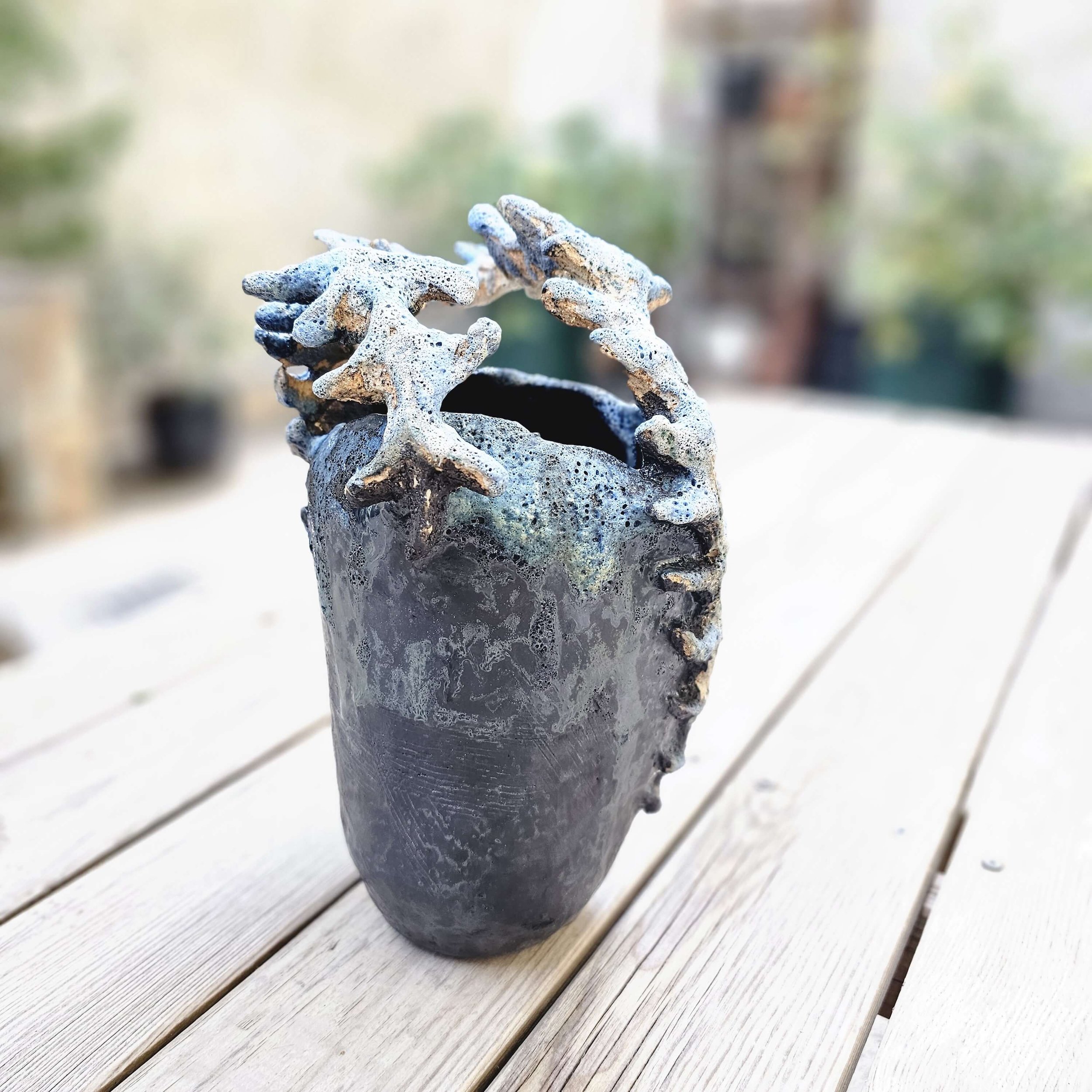 Mirsini Roumelioti. Mirsini Ceramics. Acanthus Reverie II. A black ceramic vase with textured surface and coral-like decorations on top, placed on a wooden table with a blurred outdoor background.