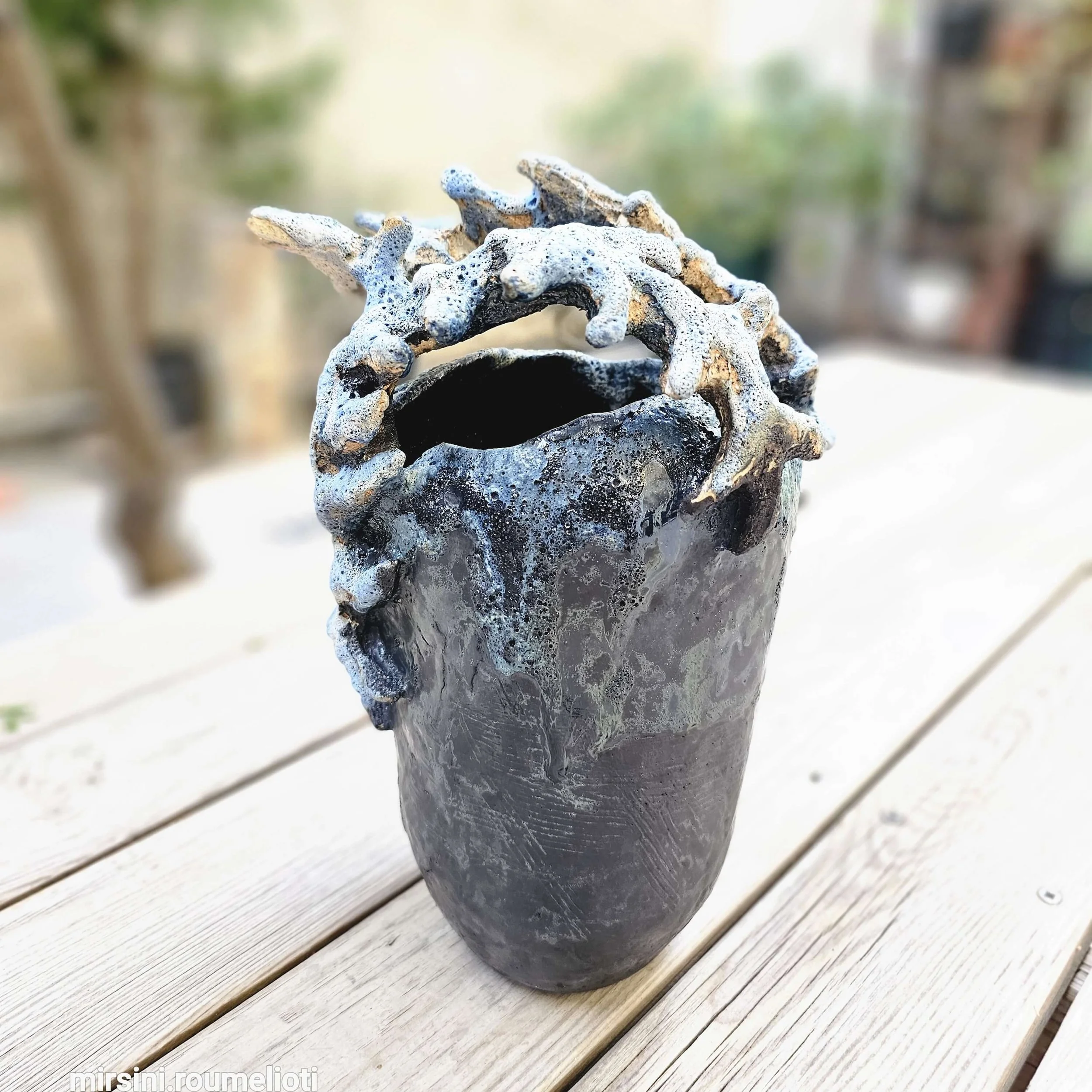 Mirsini Roumelioti. Mirsini Ceramics. Acanthus Reverie II. A ceramic vase with a textured black surface and white vine-like rim on top, placed on a wooden surface outdoors.