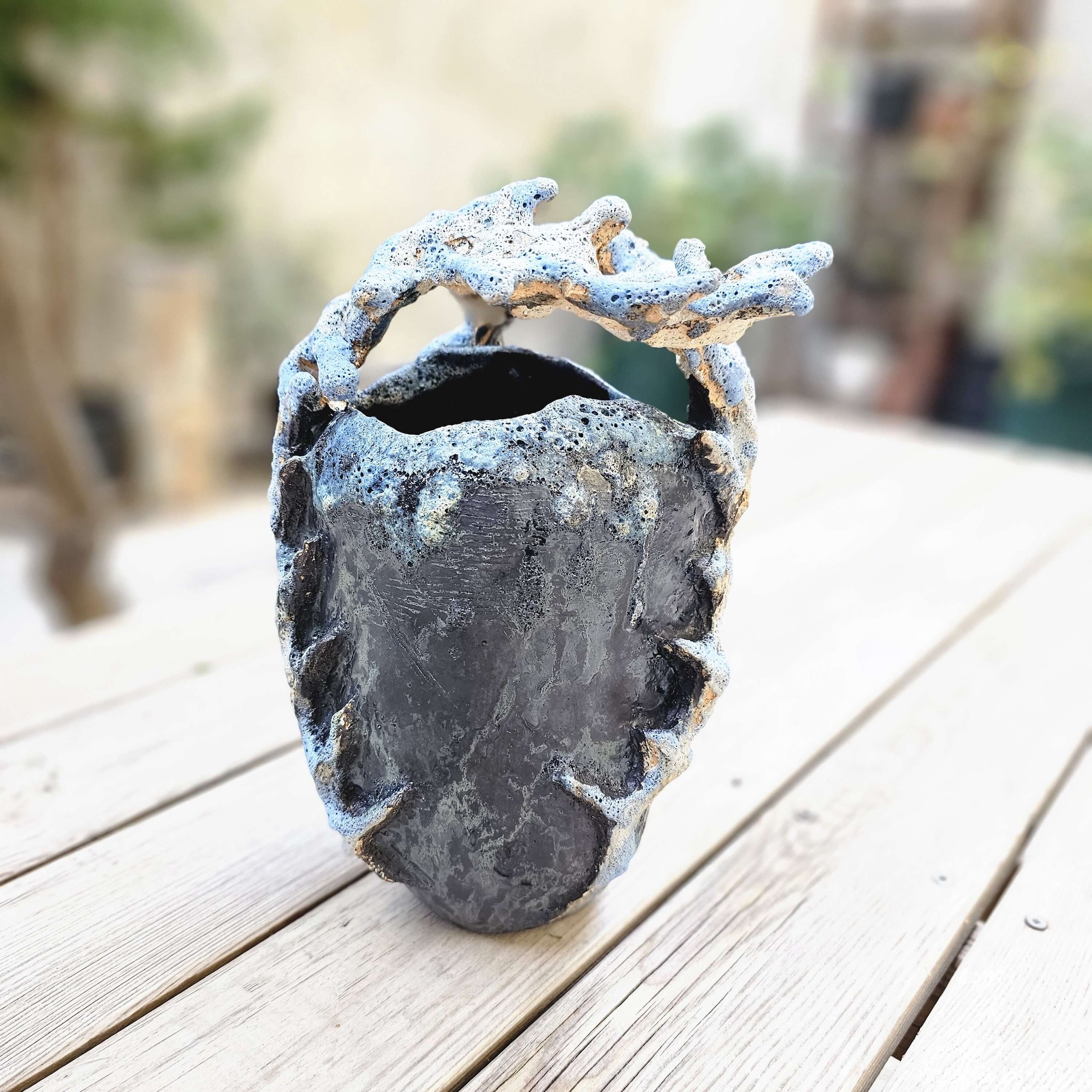 Mirsini Roumelioti. Mirsini Ceramics. Acanthus Reverie II. A ceramic vase with an irregular, textured surface, featuring a dark interior and a decorative, twisting rim, placed on a wooden surface outdoors.