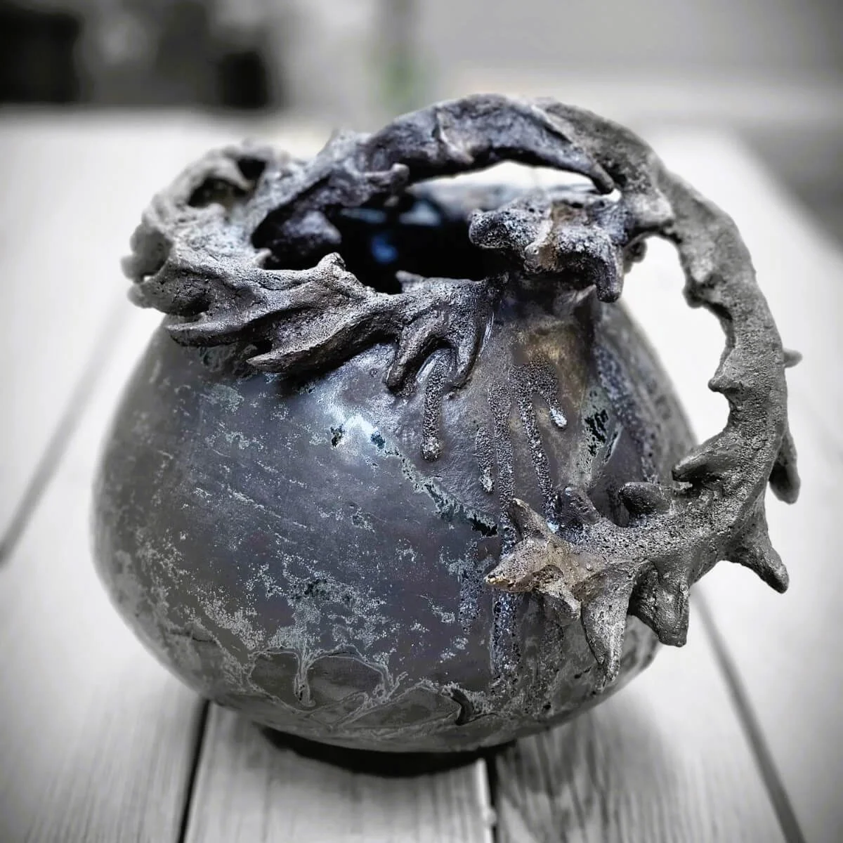 Mirsini Roumelioti. Mirsini Ceramics. Acanthus Reverie I. Sculpture of a metallic glaze ceramic vessel with a decorative leafy vine on top.