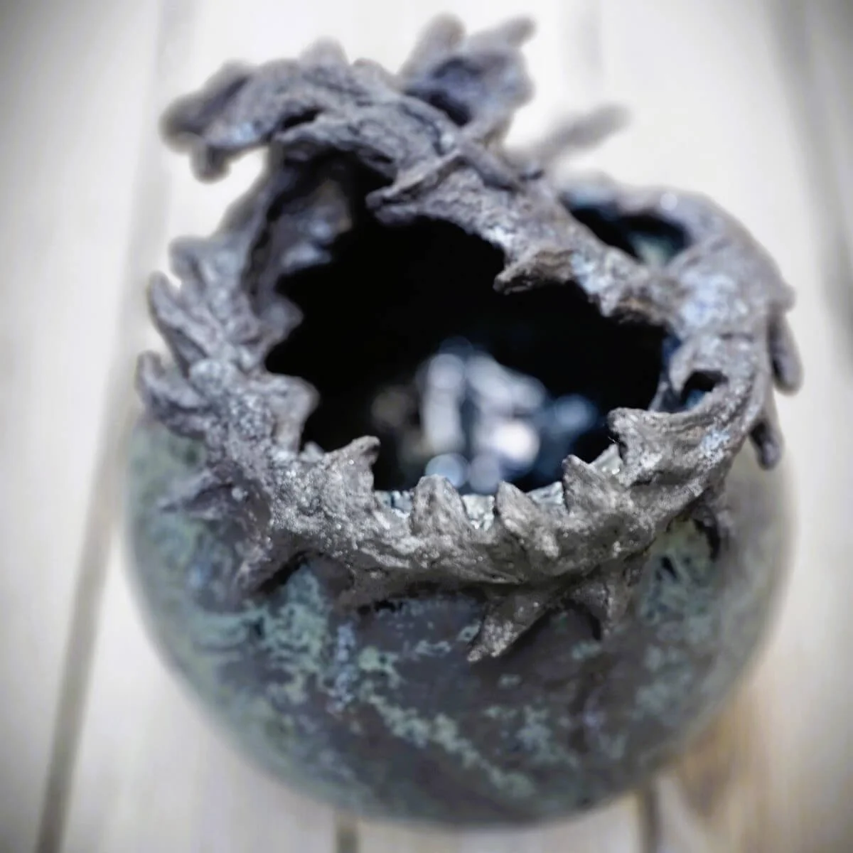 Mirsini Roumelioti. Mirsini Ceramics. Acanthus Reverie I. Sculpture of a metallic glaze ceramic vessel. Close-up of a dark, hollow, twisted metal object resembling a pine cone, with a weathered and rough texture.