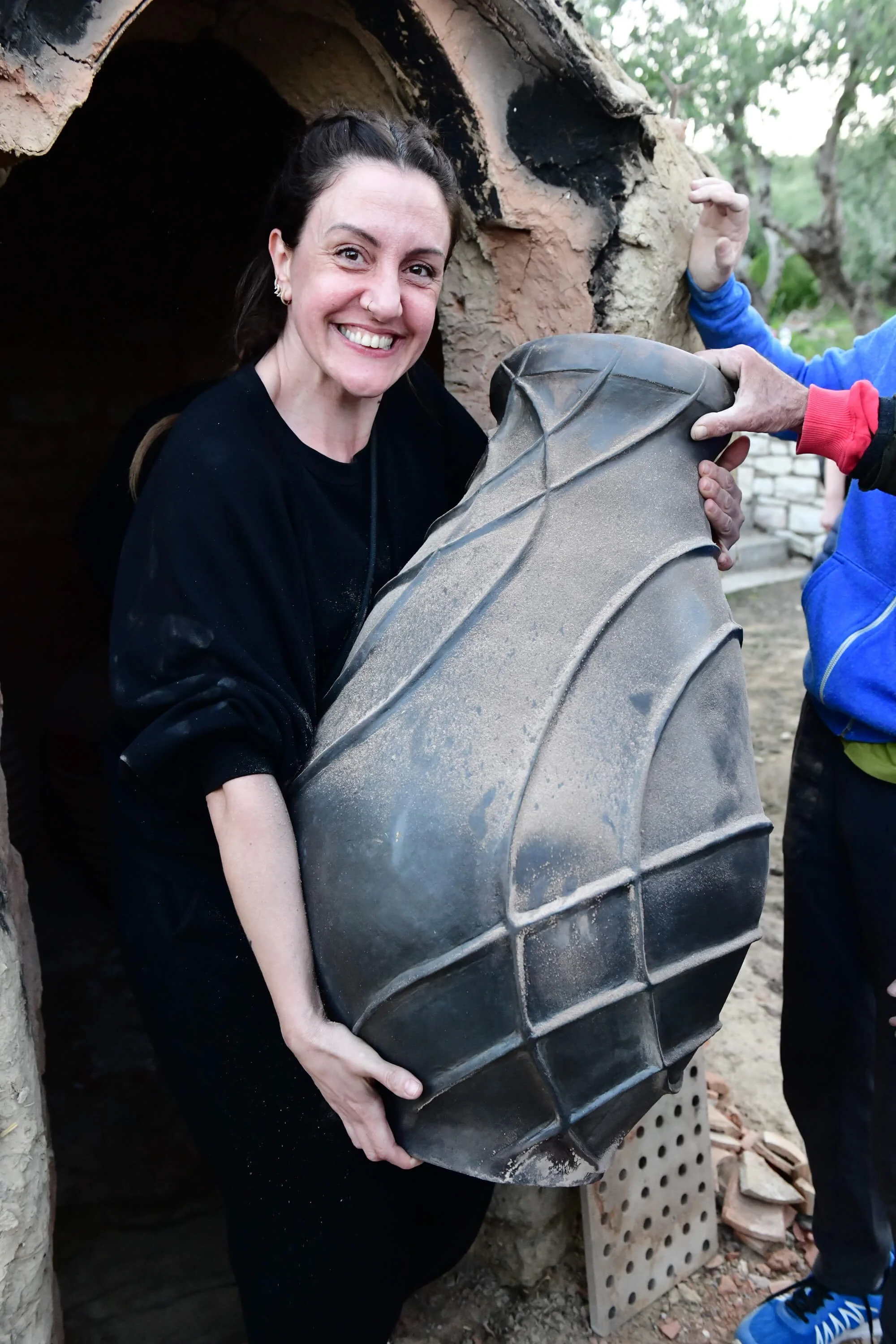 Mirsini Roumelioti holding a hand-built ceramic vessel in front of a traditional wood-fired kiln