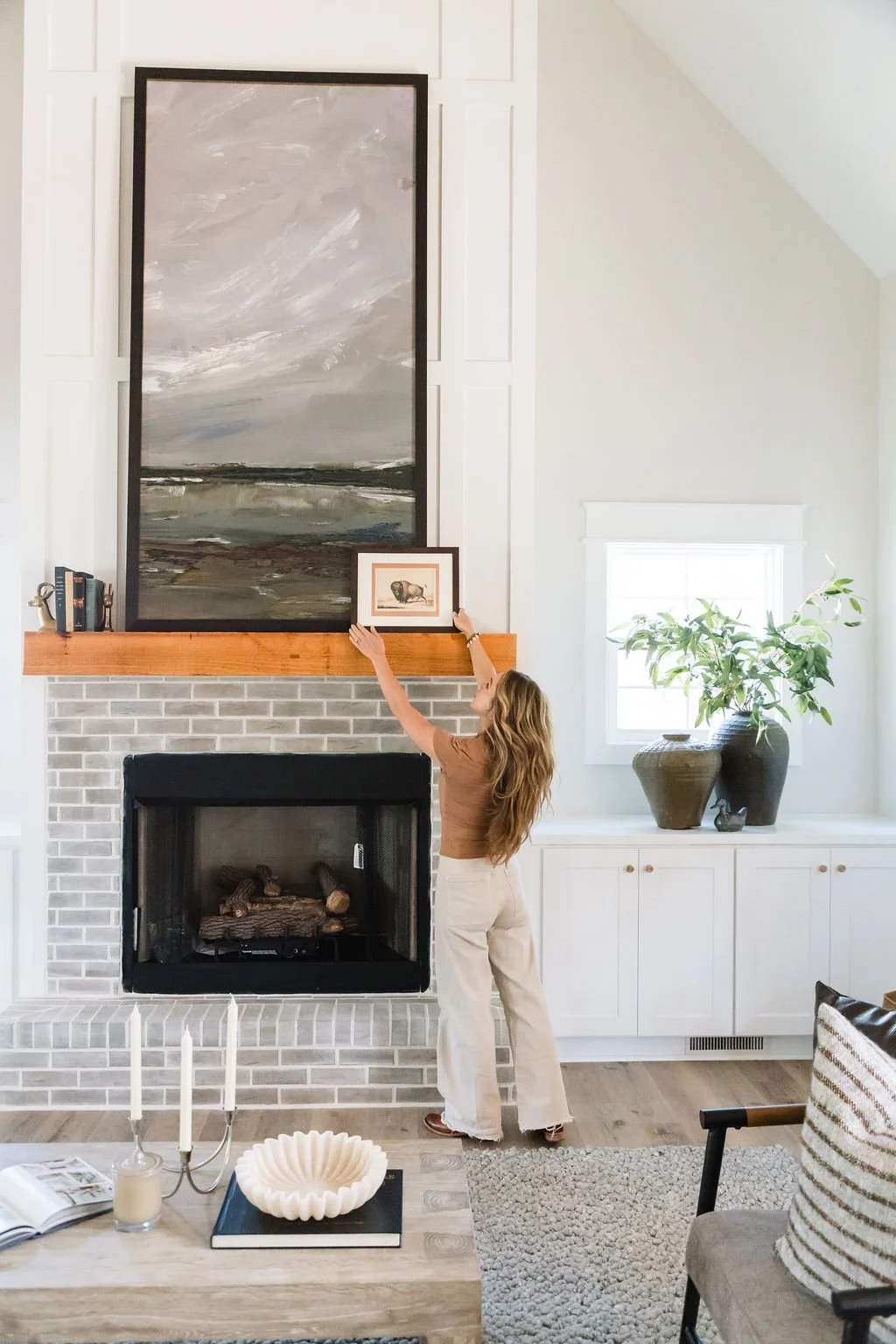 Woman hanging a framed picture above a fireplace in a living room with a large landscape painting, a sunlit window, a potted plant, and modern decor.