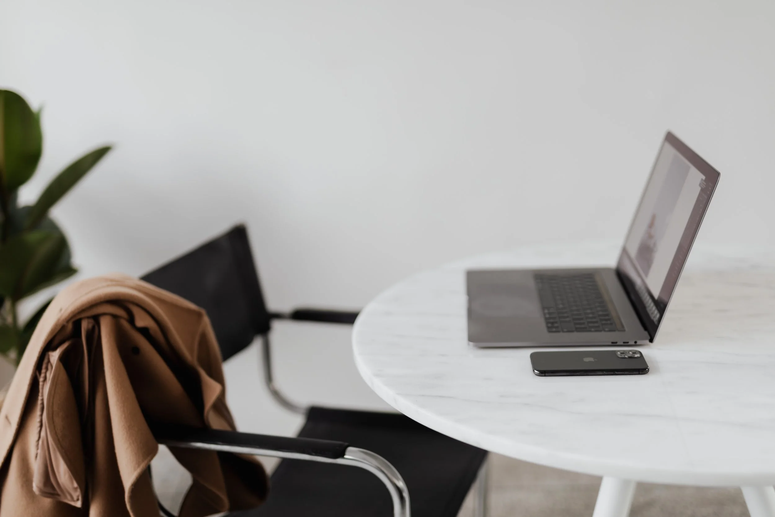 A laptop and a smartphone are placed on a white marble table in a minimalist office setting, with an empty black chair nearby and a tan coat draped over its back.