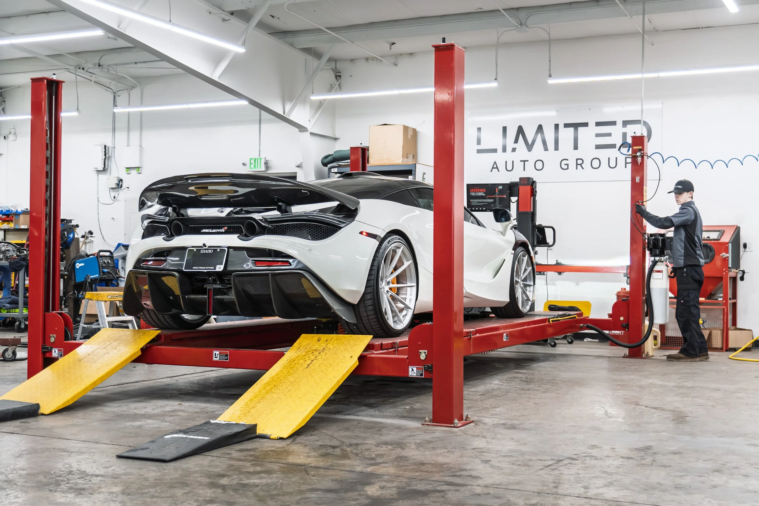 High-performance white sports car on a hydraulic lift in an auto workshop, with a person in black attire working nearby, surrounded by tools and equipment.
