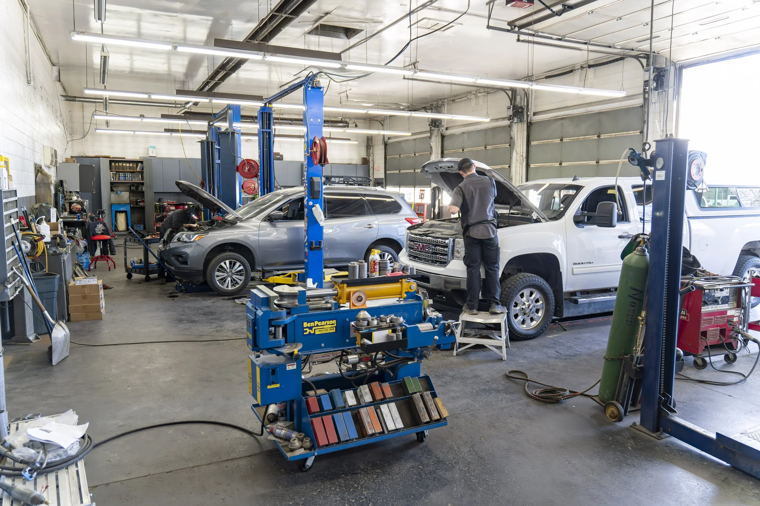 Automotive repair shop with two vehicles raised on hydraulic lifts, mechanics working on the engines, and various tools and equipment in the work area.