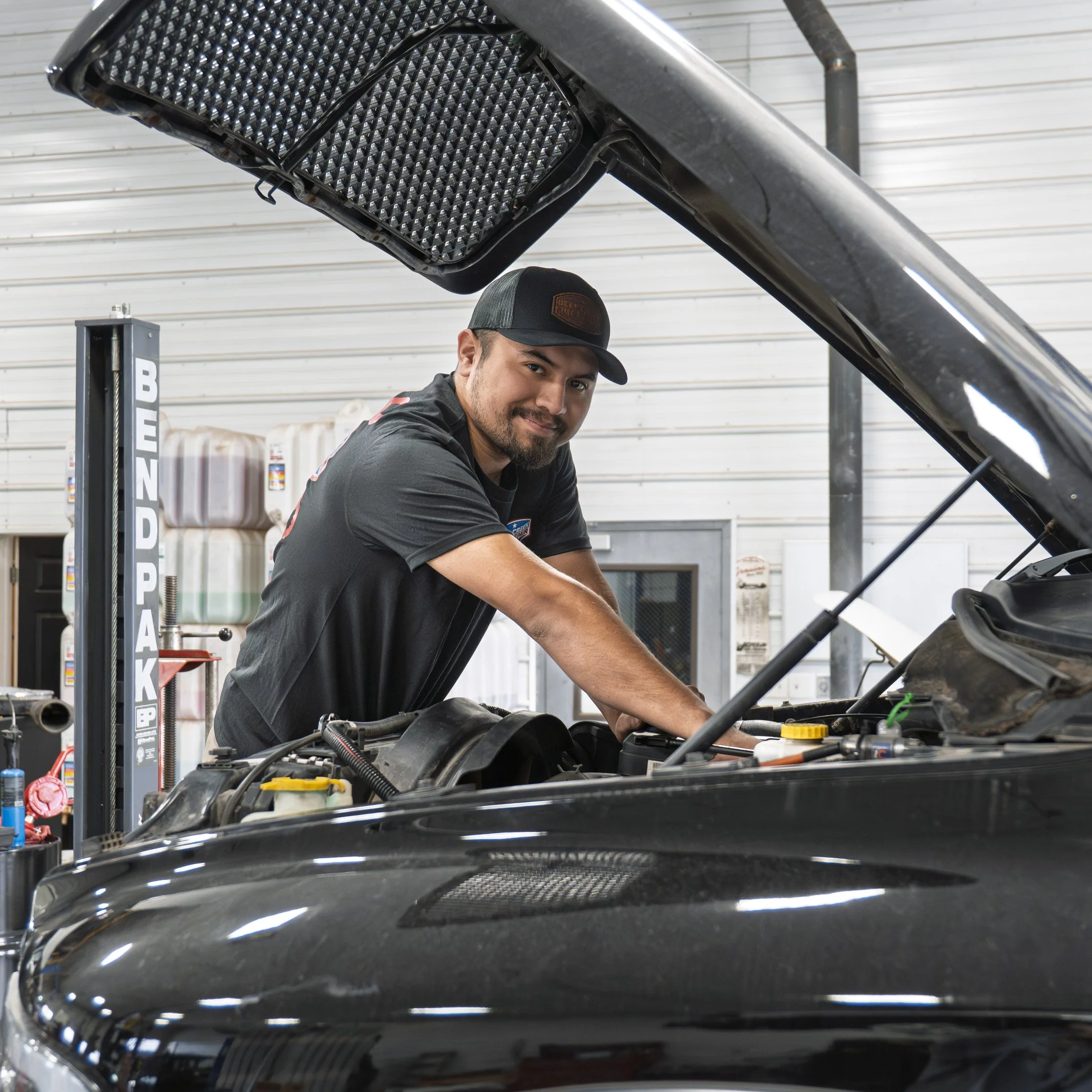 A man working on a car engine in a garage, wearing a black cap and black shirt, with a slight smile, with car hood open and workshop tools and supplies in the background.