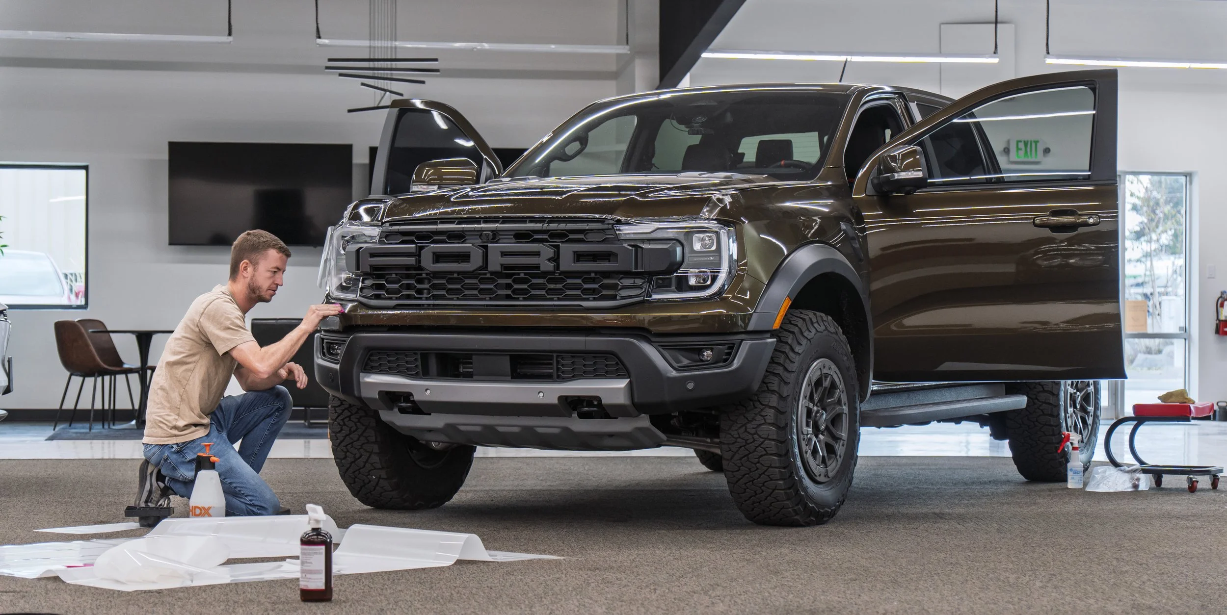 A man working on a brown Ford F-150 Lightning truck inside a showroom or service area, with various tools and cleaning supplies nearby.