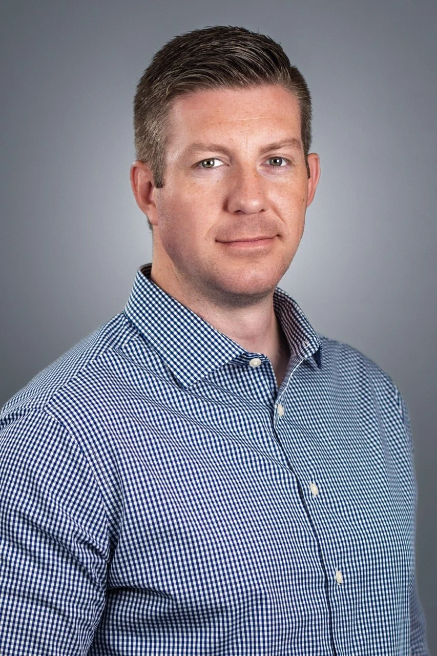 Headshot of a man with short brown hair wearing a blue and white checkered shirt against a gray background.