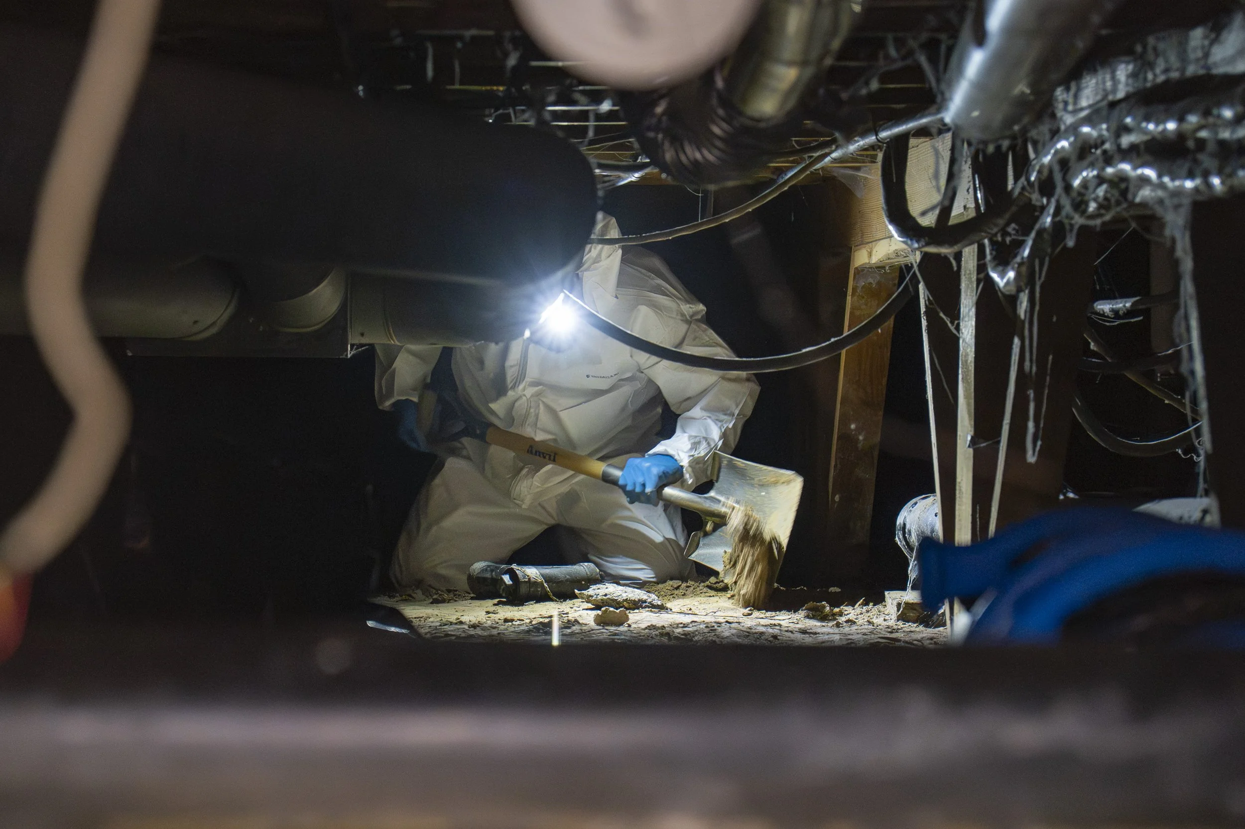 A worker in white protective gear and blue gloves is moldings a drywall with a sledgehammer while kneeling under a structure with pipes and wiring in a dark space illuminated by a flashlight.
