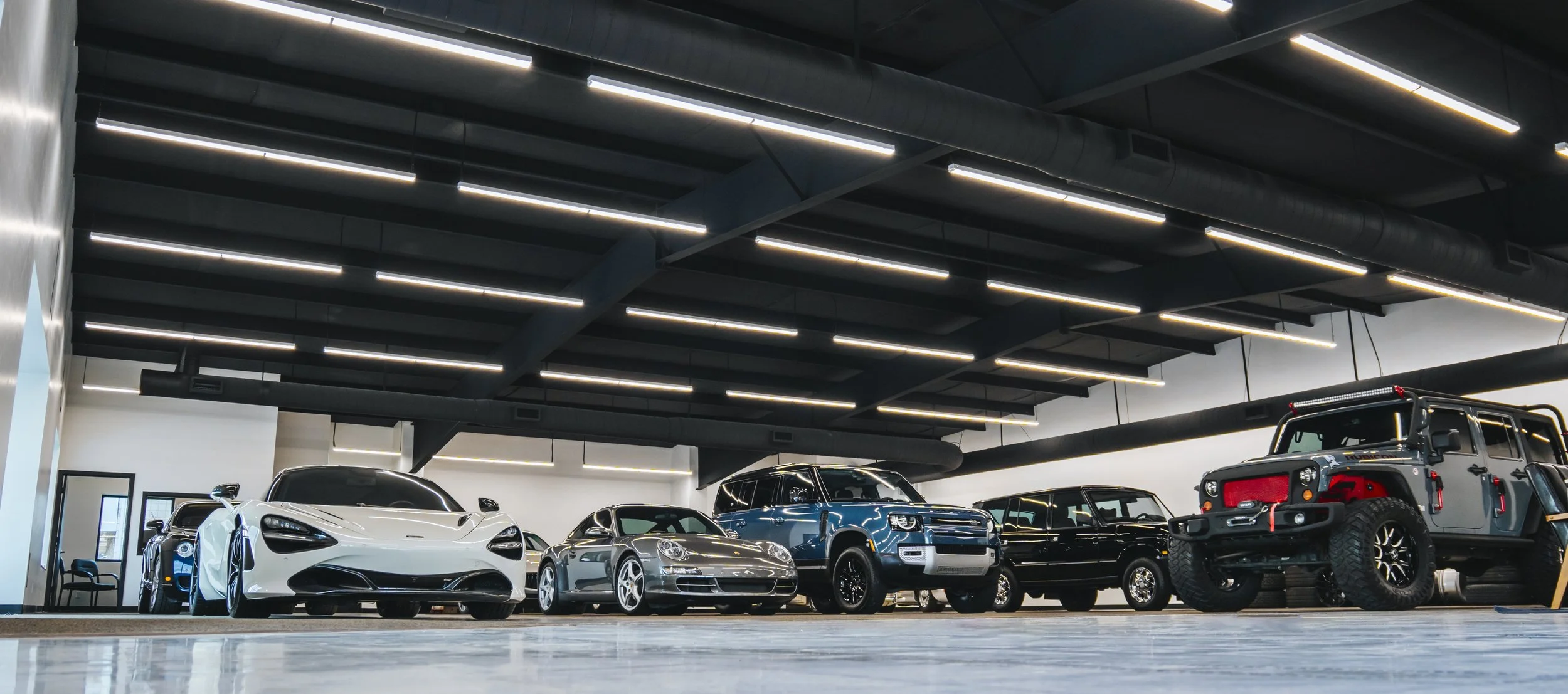 Inside a modern car showroom featuring several luxury vehicles including a white sports car, silver sedan, metallic blue SUV, black luxury car, and an off-road Jeep, under a dark ceiling with linear lighting.