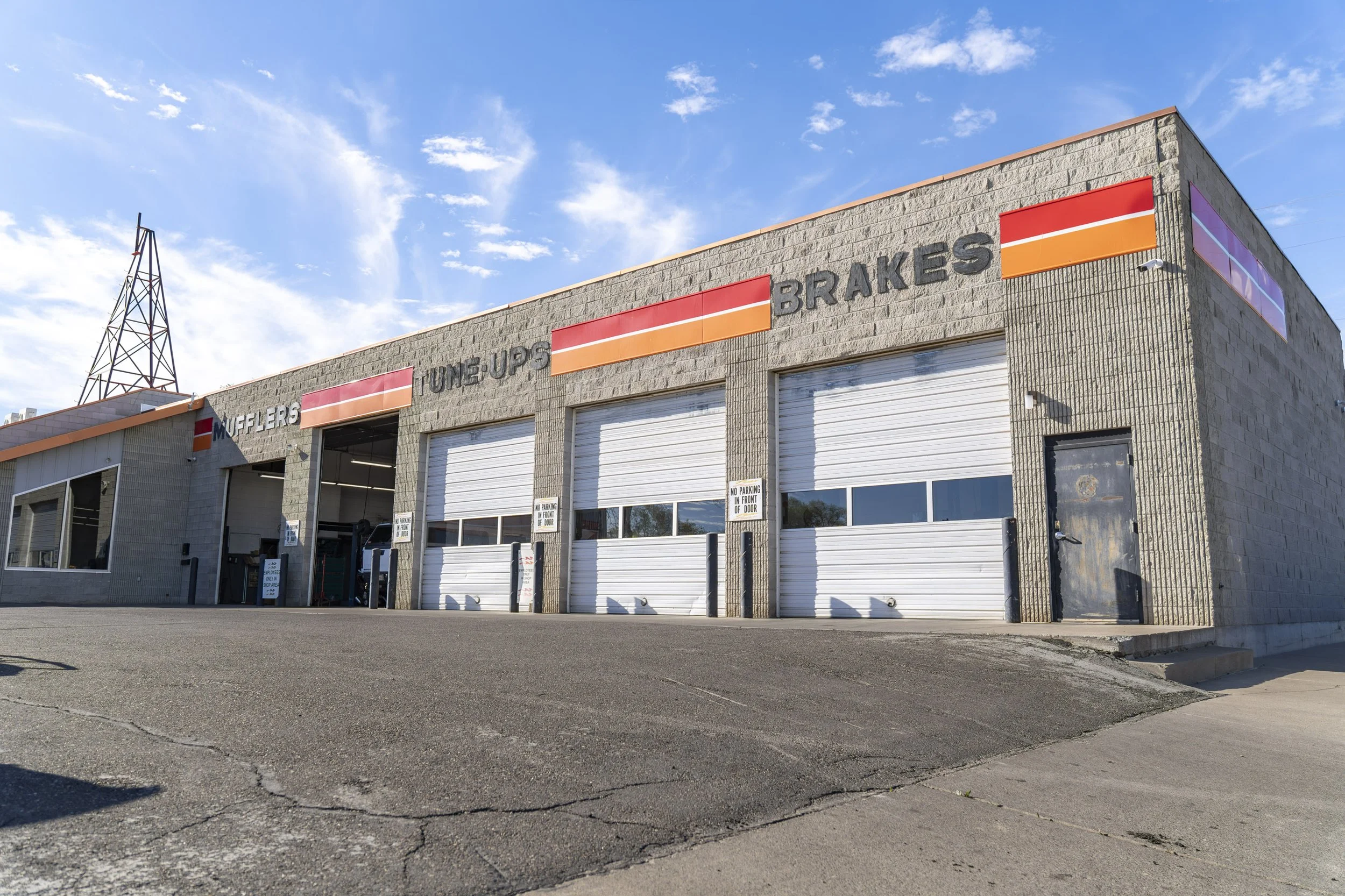 Automotive repair shop with garage doors and signs for mufflers, tune-ups, brakes, against a blue sky with clouds.