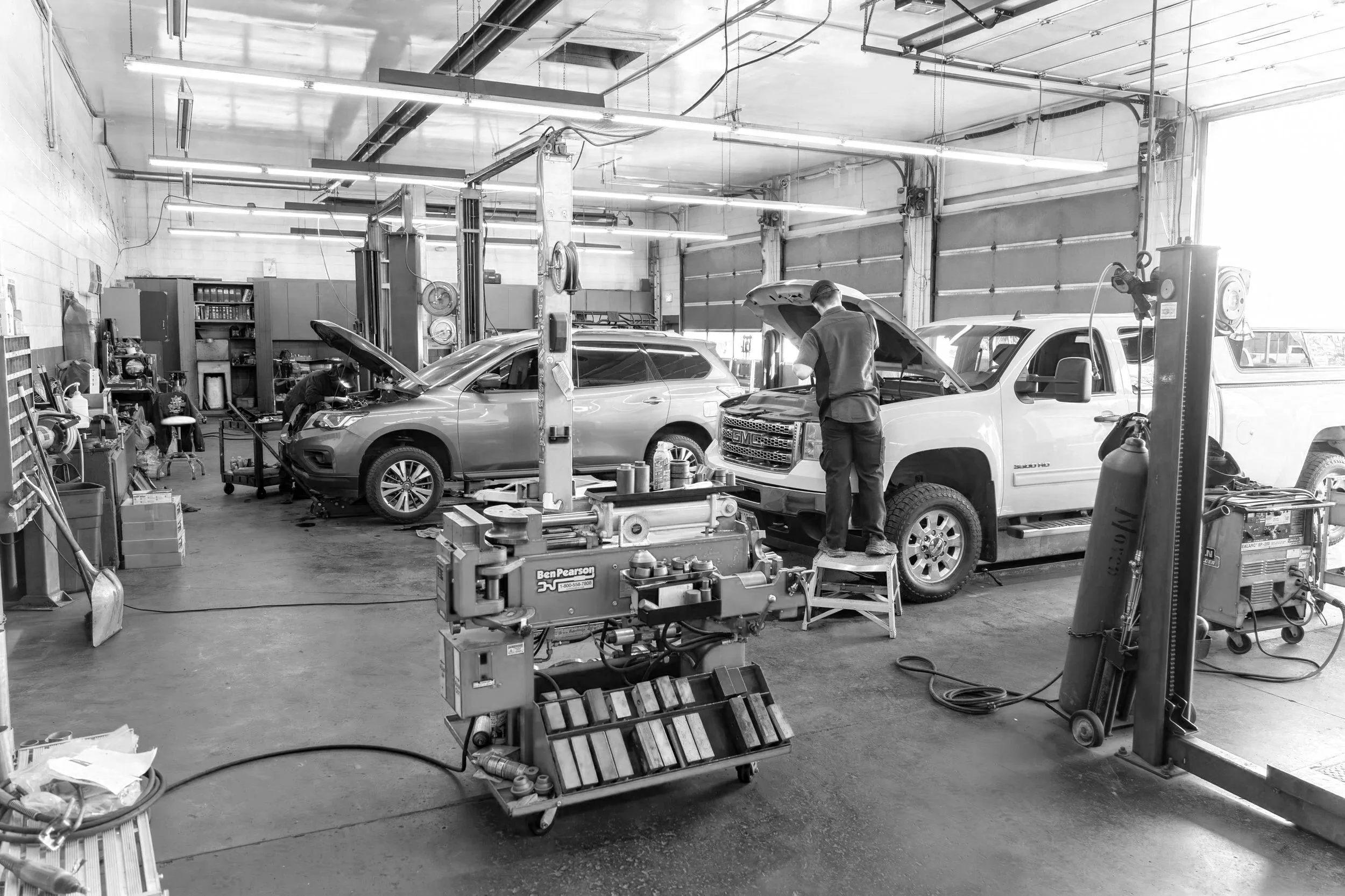 Inside an auto repair shop with two vehicles being serviced. A mechanic working on a white pickup truck with a raised hood, another person working on a car in the background. Various tools and equipment are visible around the shop.