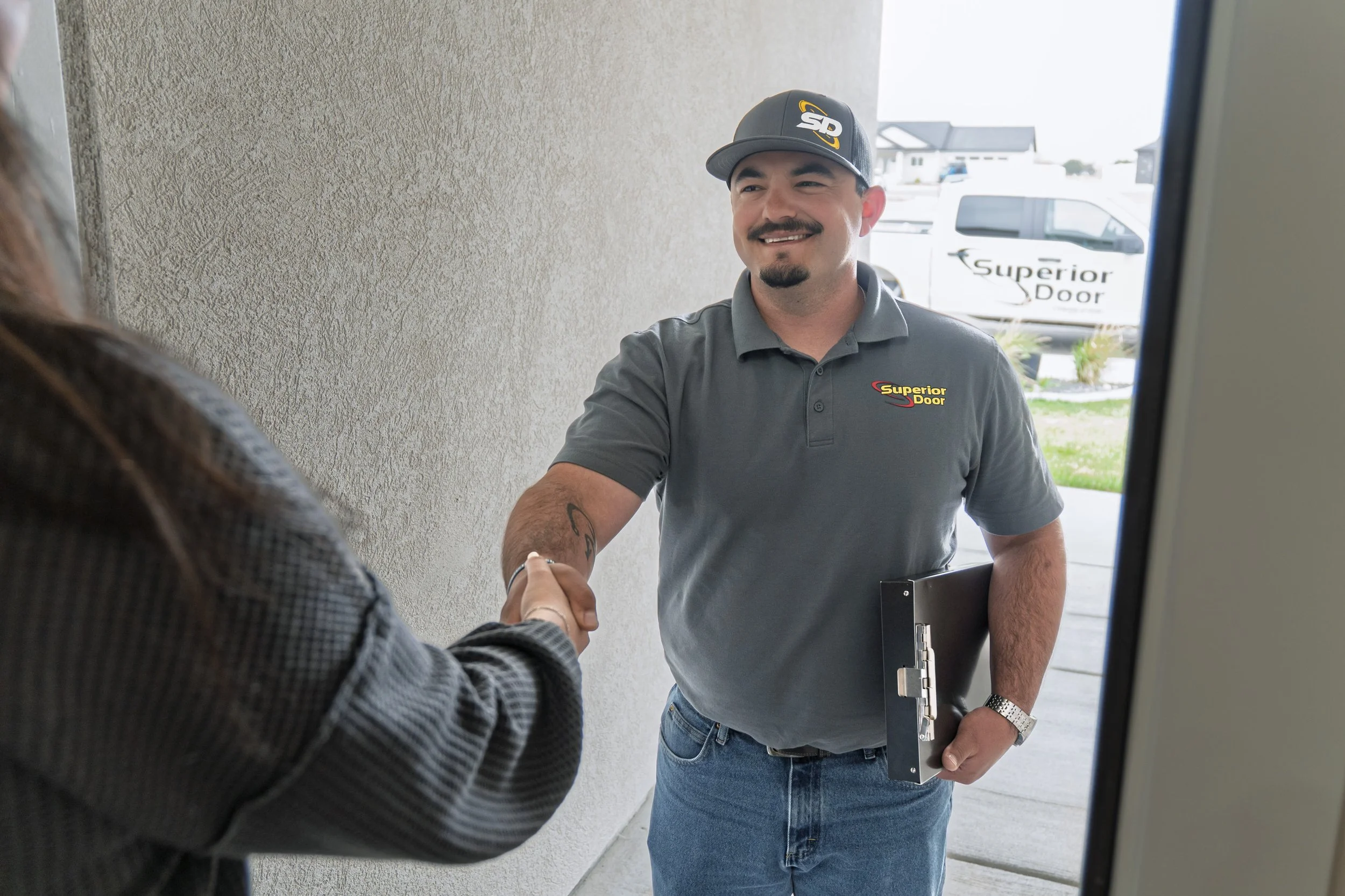 A man with a mustache and goatee, wearing a gray Superior Door shirt and cap, is smiling and shaking hands with a woman at the door. The man holds a clipboard and is outside a house in a residential neighborhood.