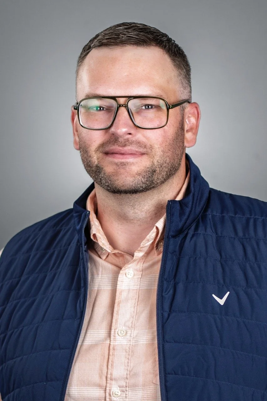 Portrait of a man with short dark hair, glasses, and a beard, wearing a peach button-up shirt and a navy blue quilted vest against a gray background.