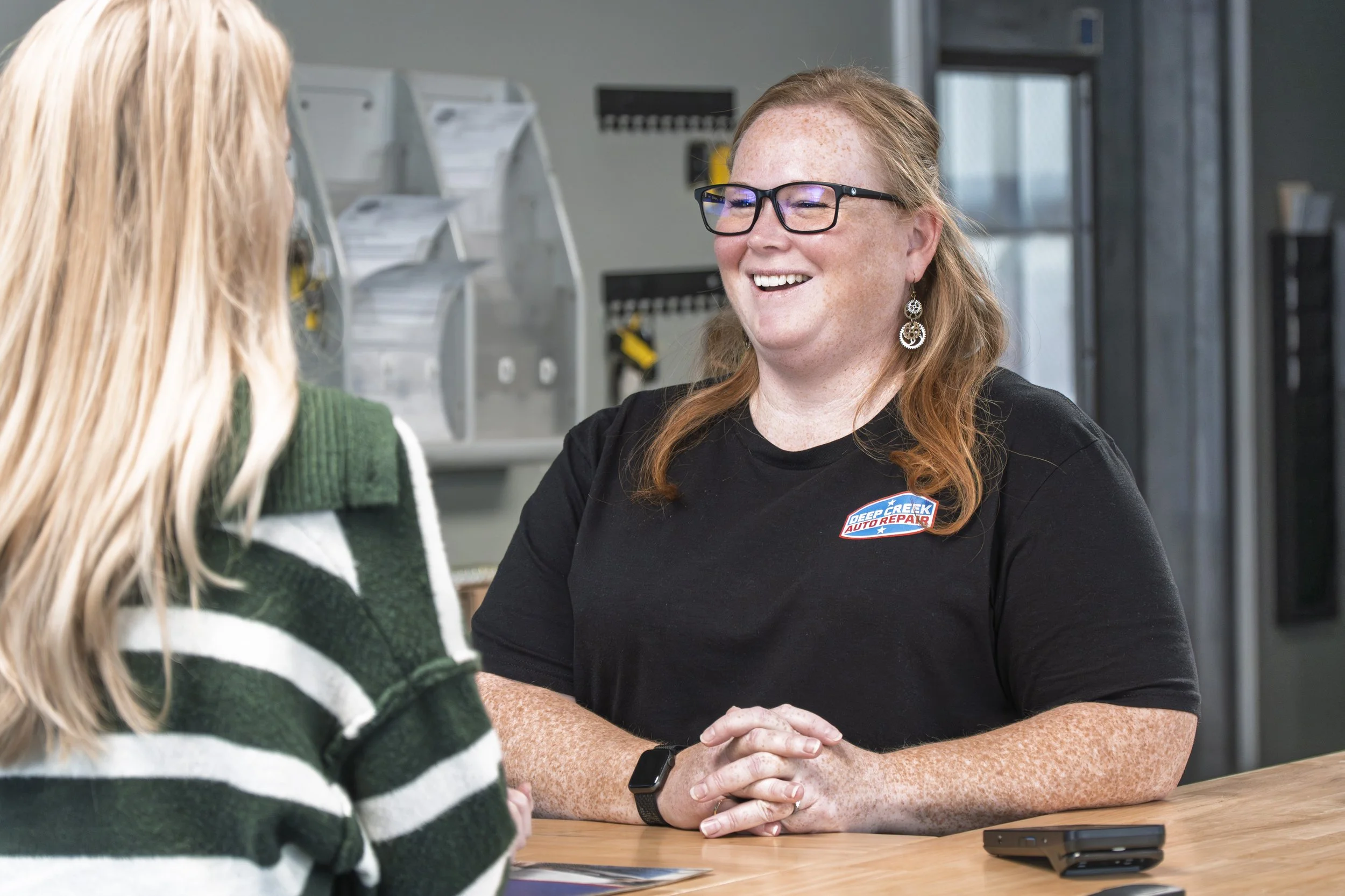 A woman with glasses, red hair, and earrings is smiling and talking to a customer at a reception desk in an auto repair shop.