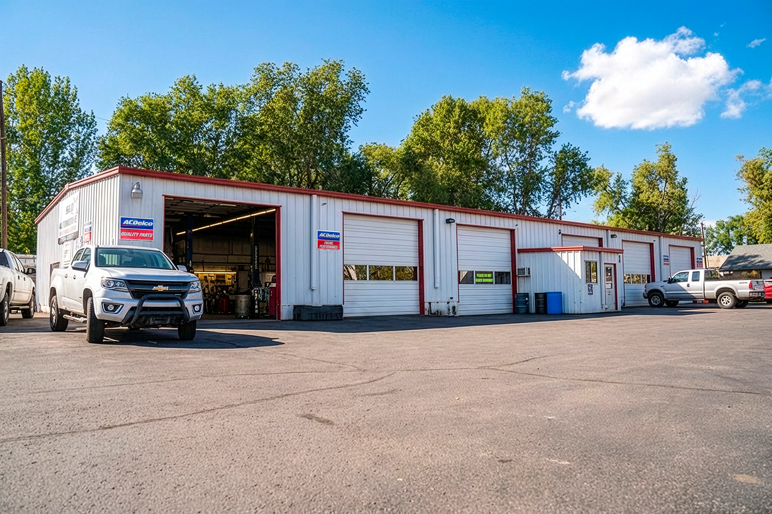 Auto repair shop with multiple bays and vehicles parked outside, surrounded by trees under a blue sky.