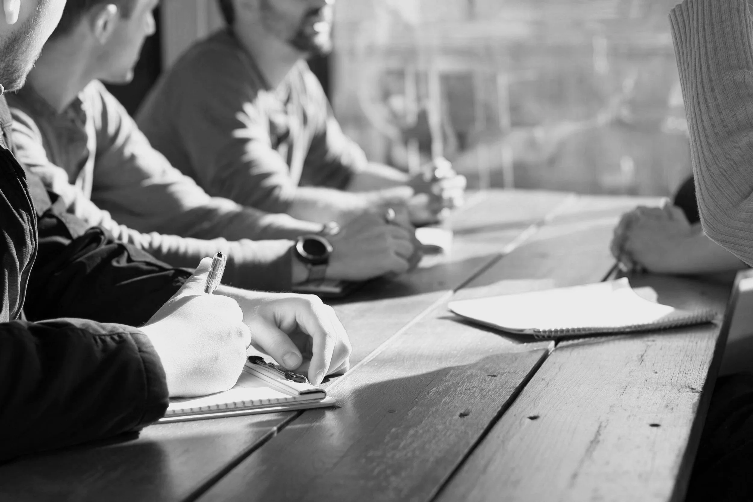 People sitting at a wooden table, taking notes, with notebooks and pens, in an outdoor setting.