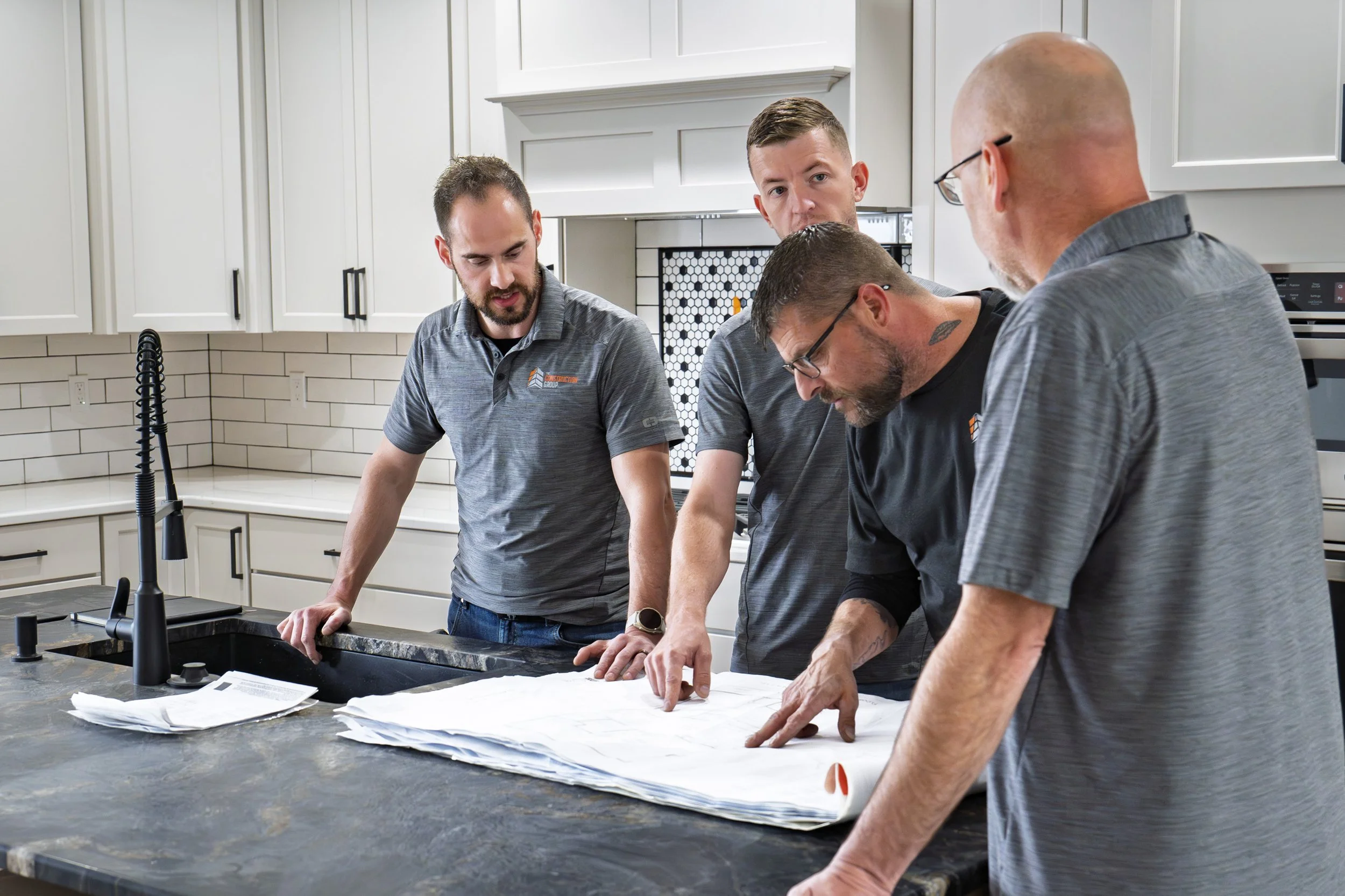 Four men wearing matching gray polo shirts gathered around a kitchen counter, examining safety plans or blueprints together.