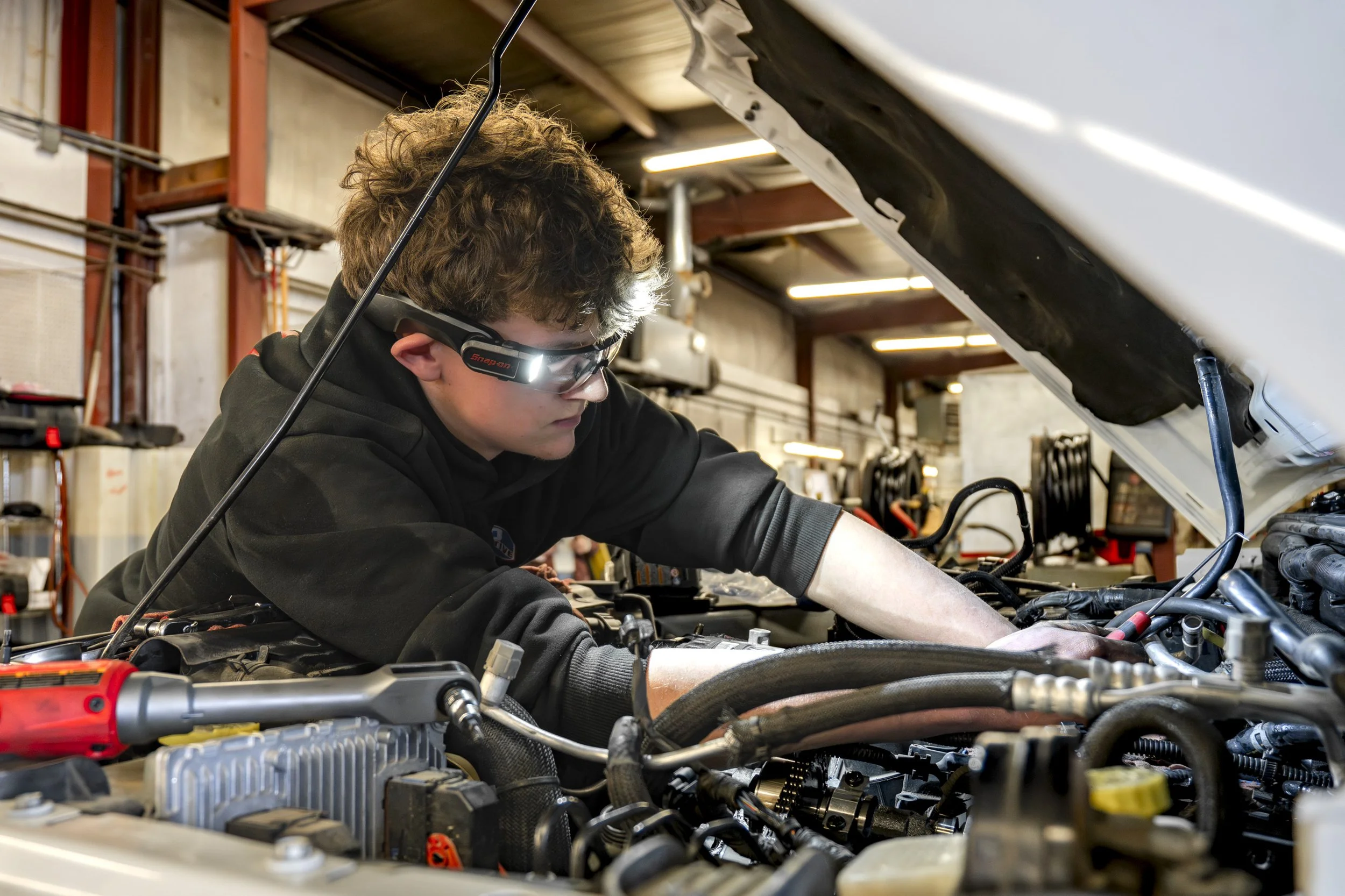 A young person working on a car engine in a garage, wearing a black hoodie and protective glasses, with the car's hood open.