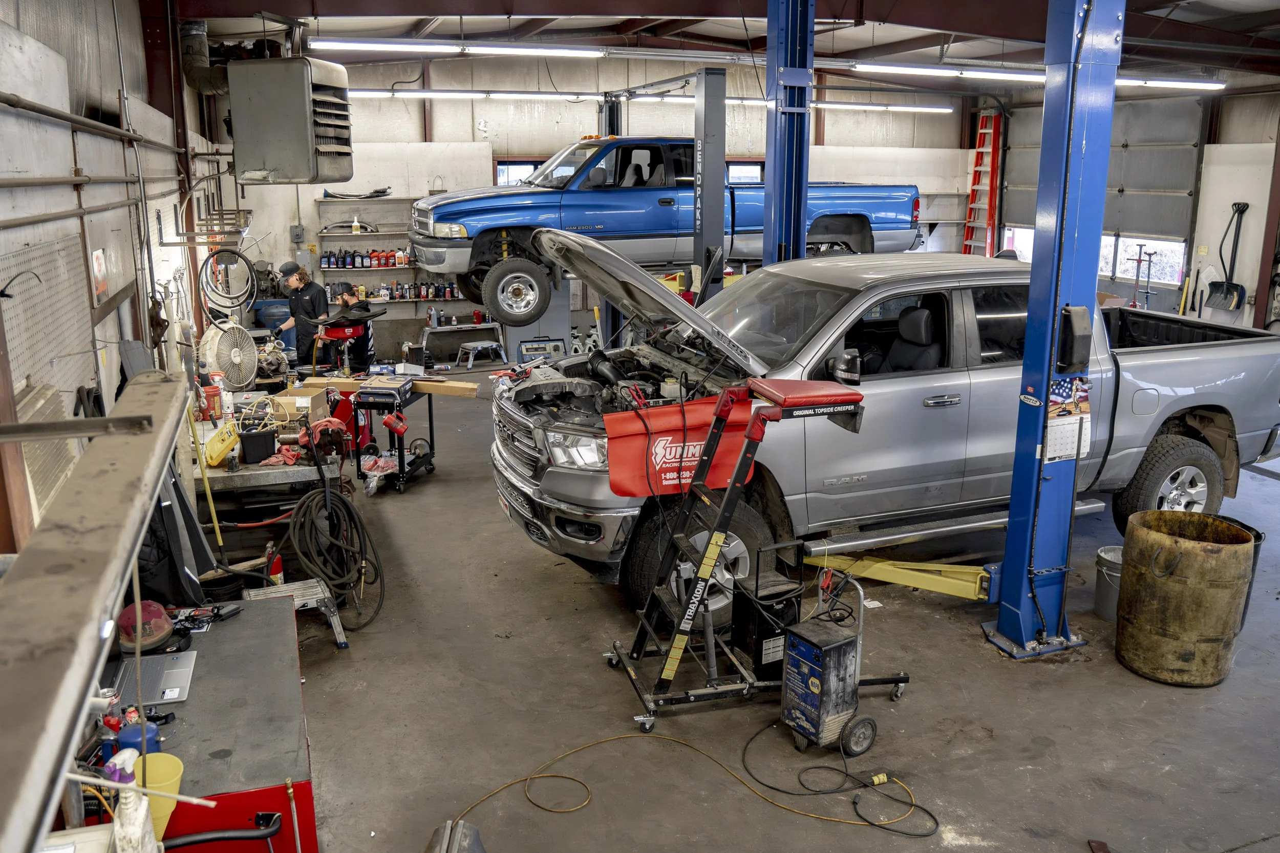 An automotive repair shop with two vehicles being serviced, one on a lift and the other on the ground, surrounded by tools and equipment.