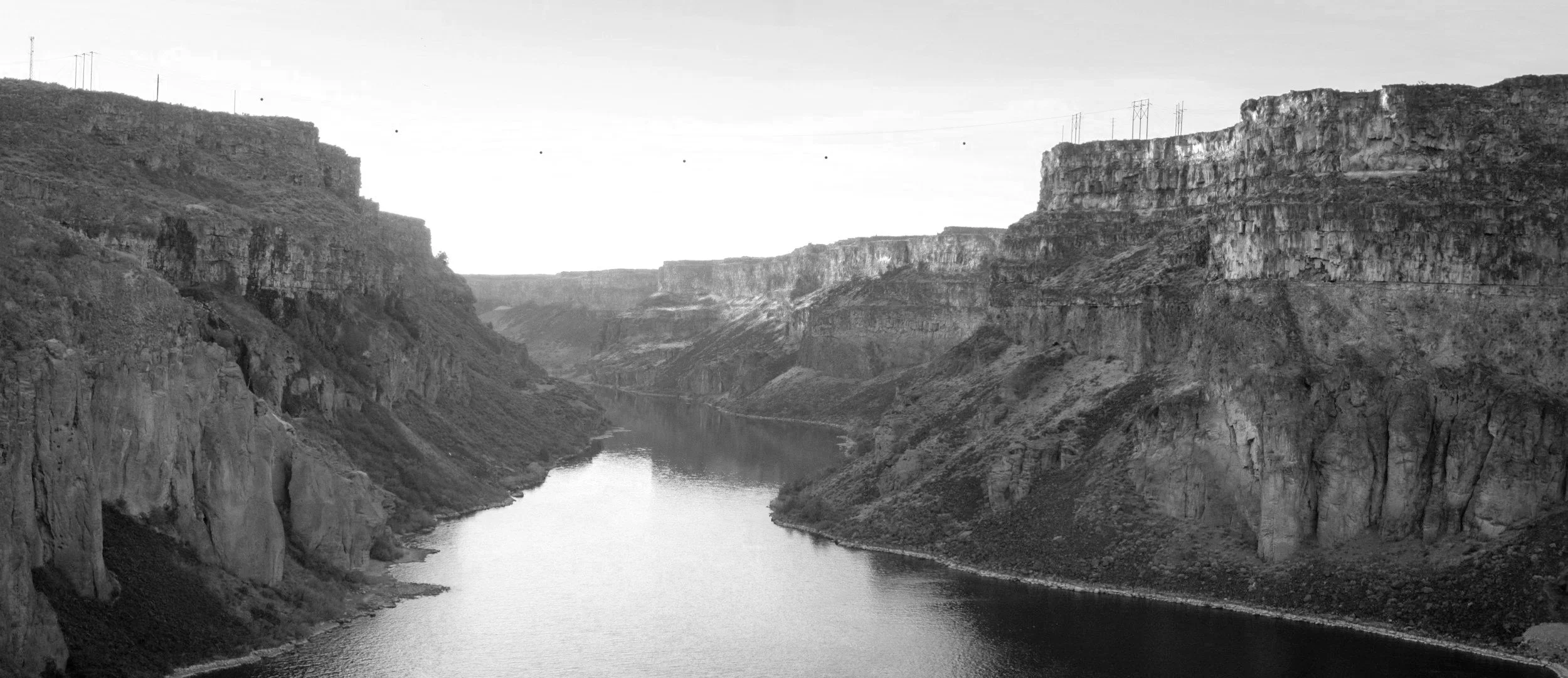 Black and white photo of a river running through a canyon with high rocky cliffs on either side. Power lines are visible above the cliffs.