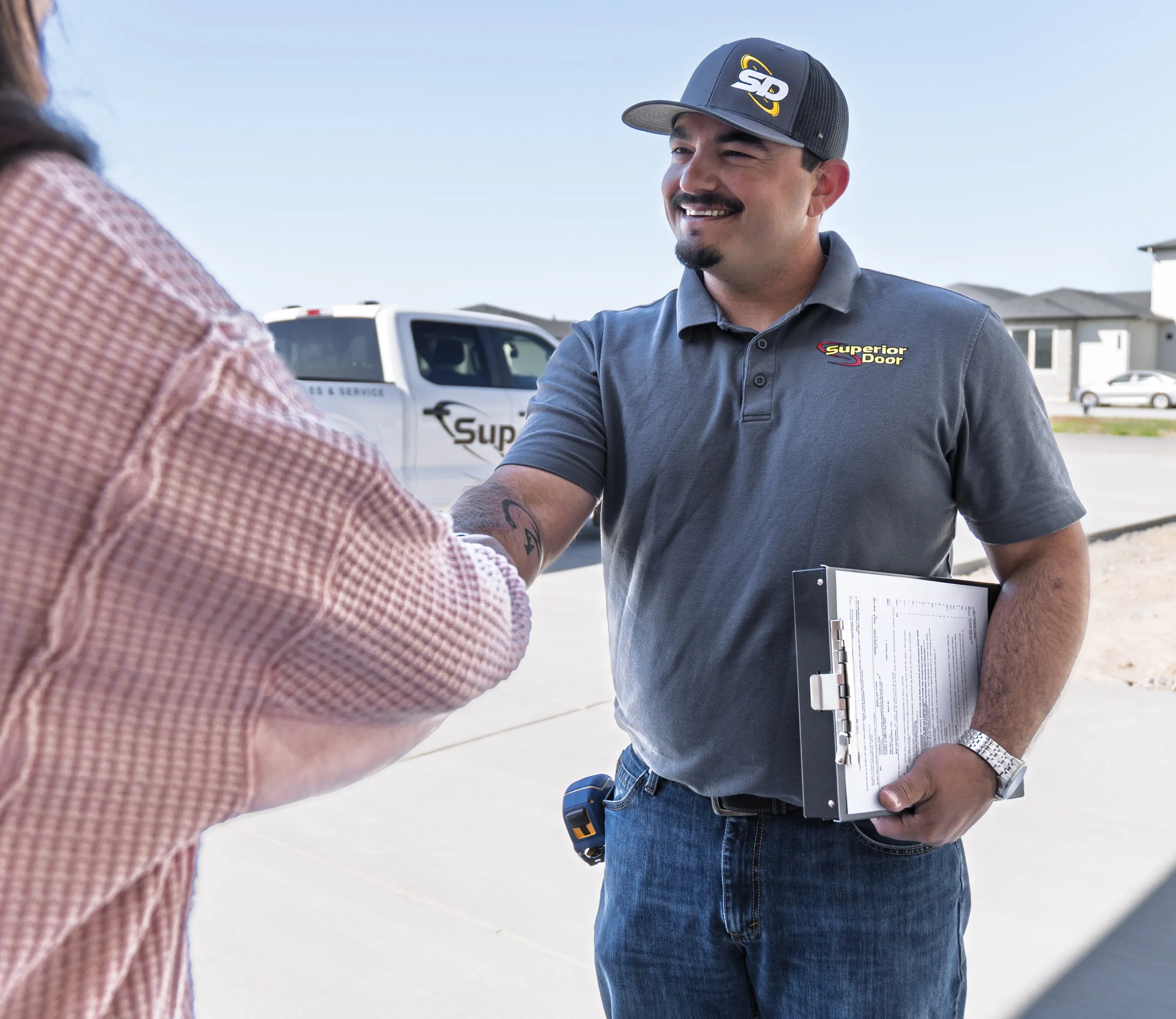A man in a gray polo shirt and a baseball cap shaking hands with a woman in a pink checkered shirt outdoors. The man is holding a clipboard and is smiling, with a vehicle in the background.