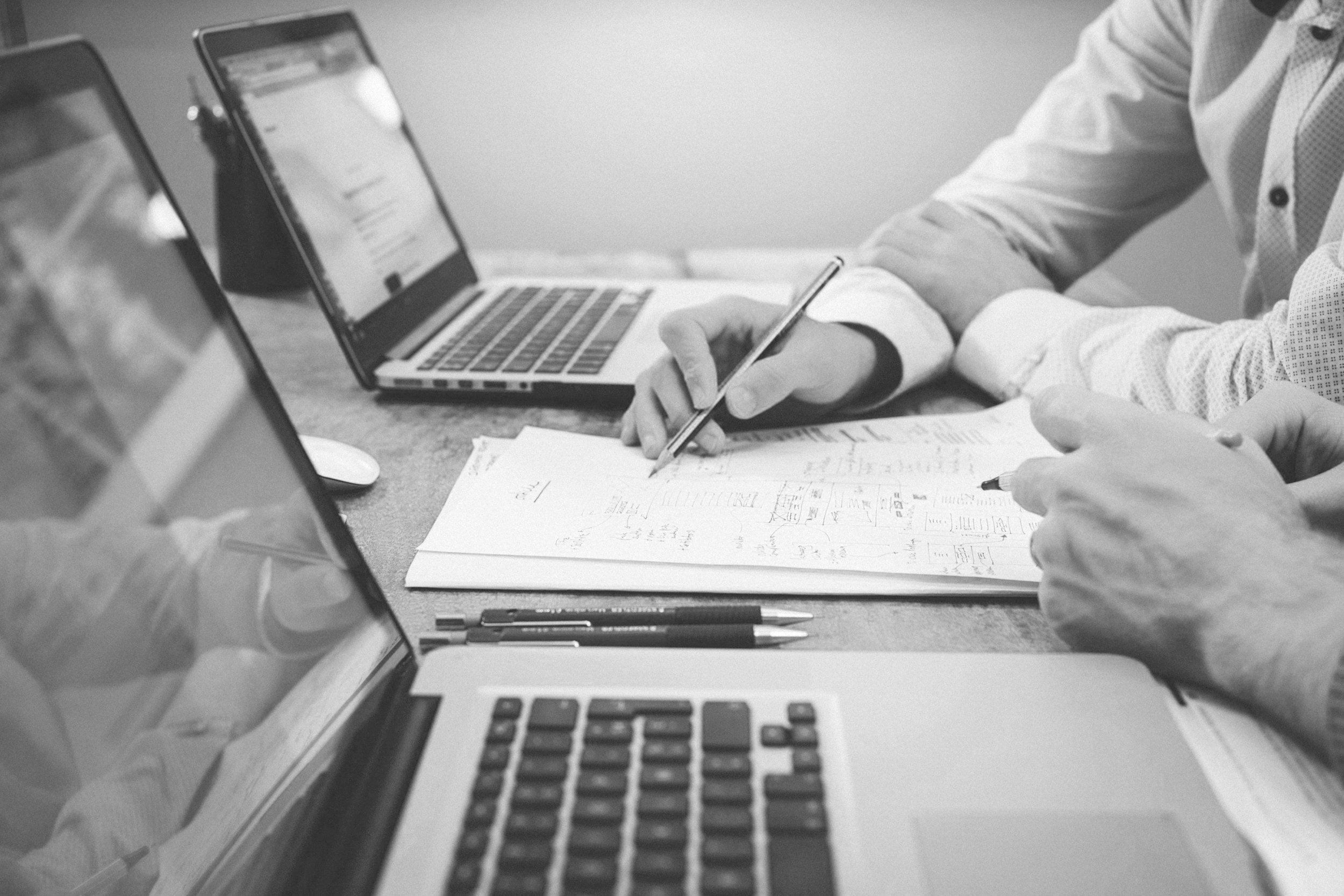 Two people working at a desk with open laptops, notebooks, and pens, one person writing on a paper.