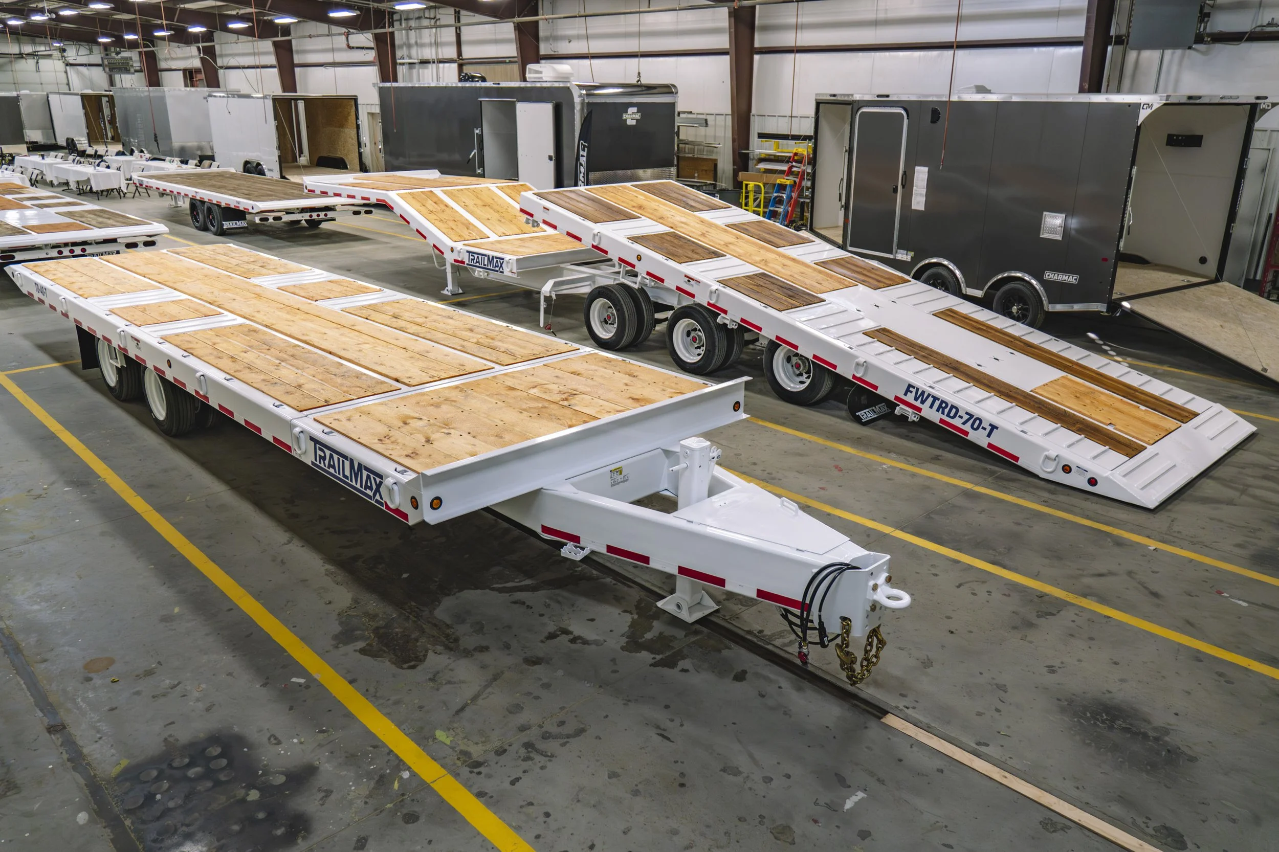 Inside a warehouse with multiple flatbed trailers and enclosed cargo trailers, some with ramps, all parked on concrete floor with yellow parking lines.