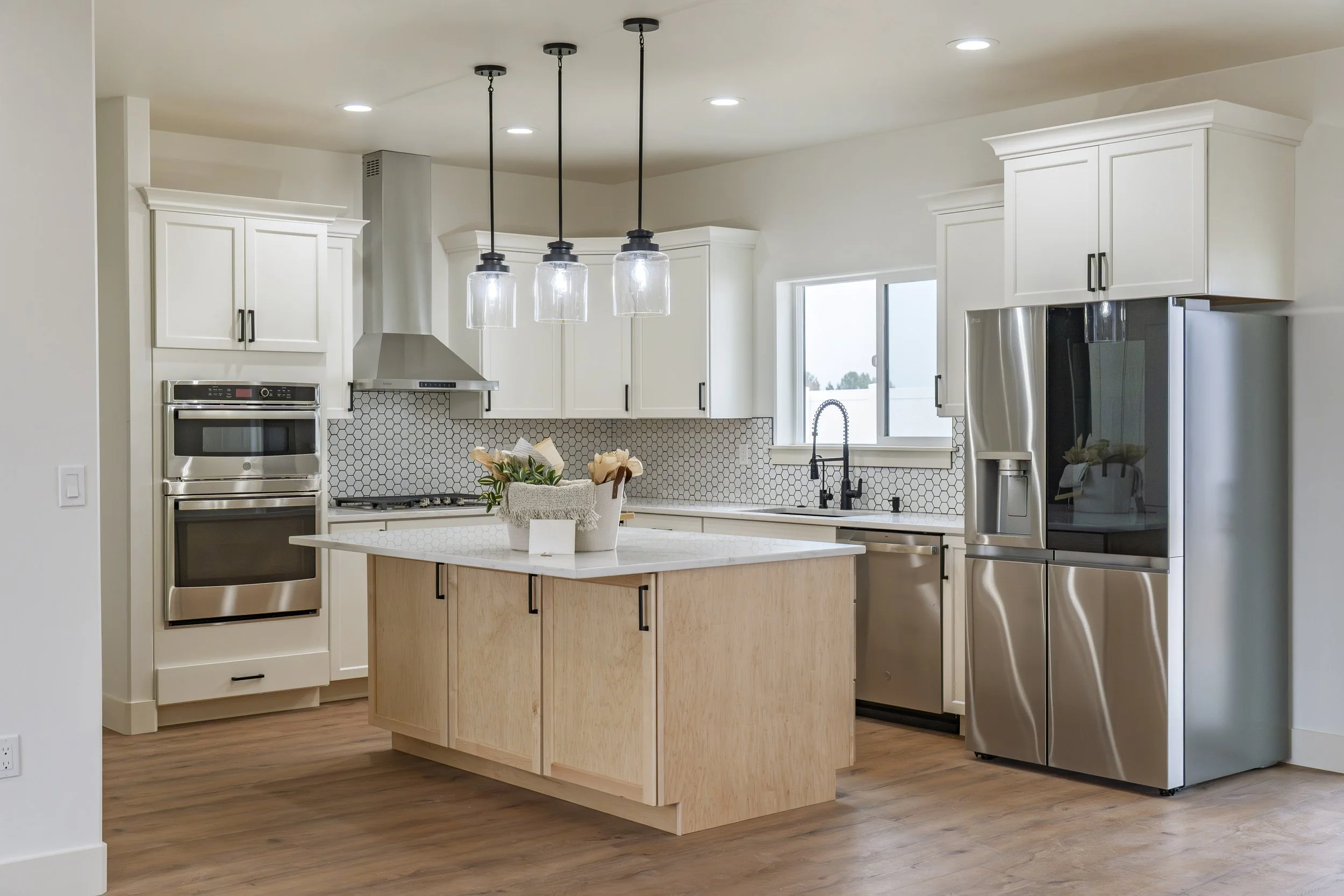Modern kitchen with white cabinets, stainless steel refrigerator, built-in oven, and island with a white countertop and a flower arrangement. Pendant lights hang above the island.