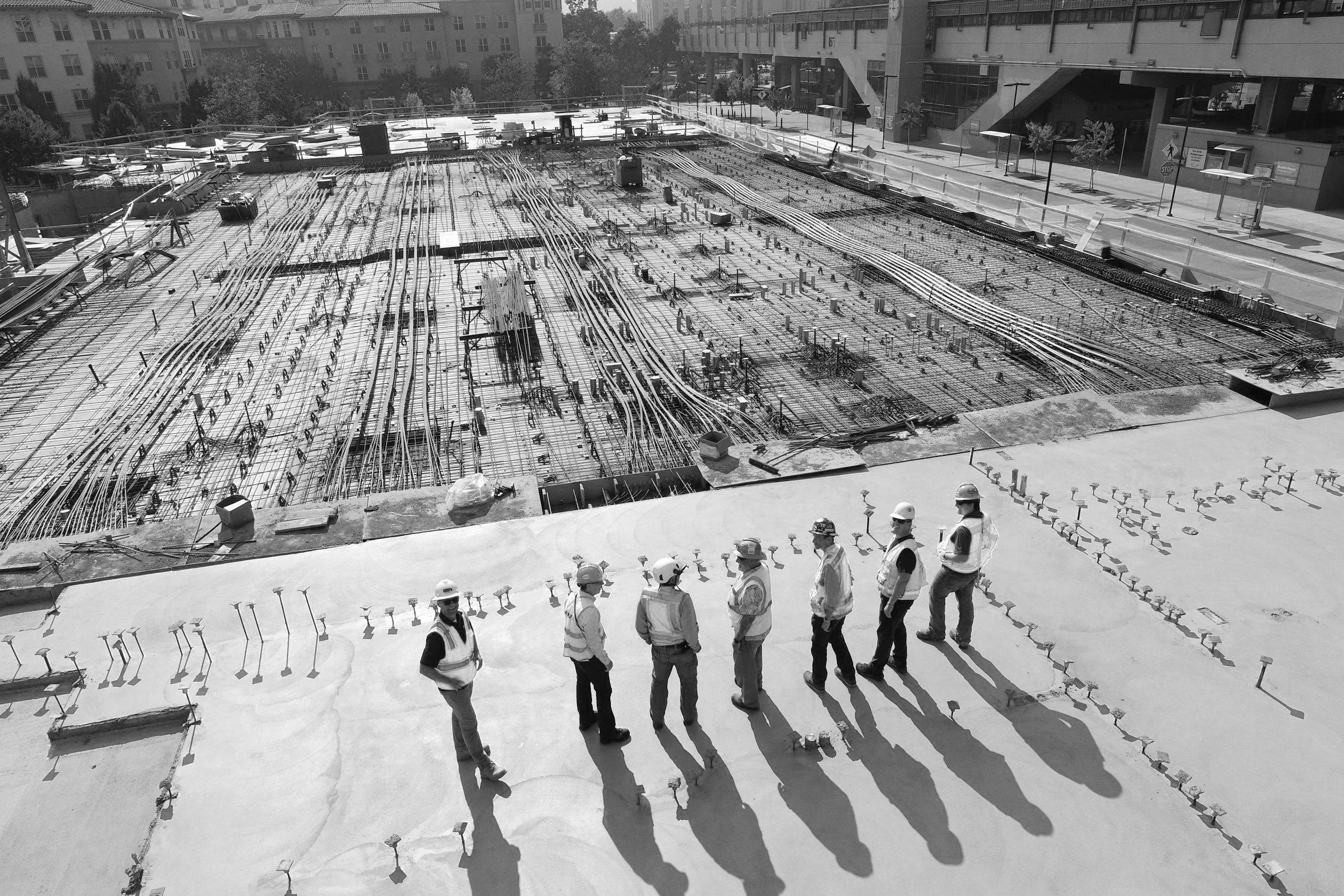 Construction site with workers wearing safety gear standing on a concrete slab, overlooking an area with steel rebar and construction materials.